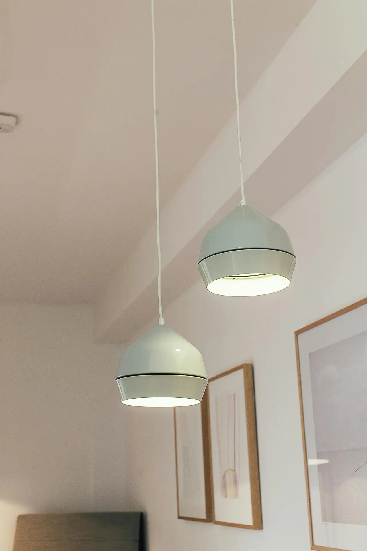 Two pastel finish pendant lamps with a soft mint green hue hang from the ceiling at different heights, casting a warm light downward. The rounded, two-tiered shades offer a clean and stylish representation of mid century modern lighting, complemented by minimalist framed art in the background.