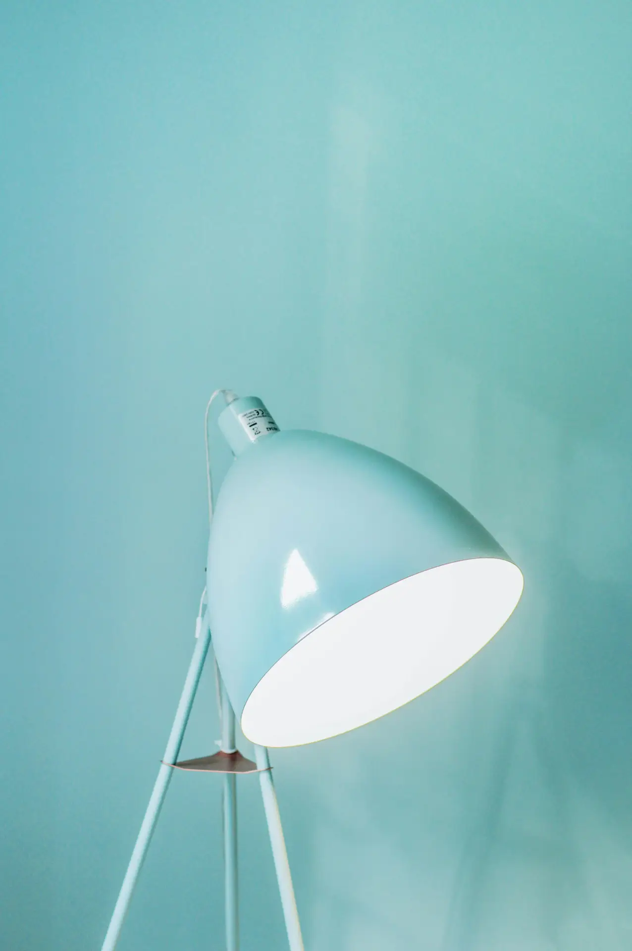 A stylish retro-look tripod floor lamp in a soft light blue finish, featuring a large adjustable metal dome head that casts a bright white light. Positioned against a matching turquoise wall, the minimalist three-legged stand exemplifies sleek mid century modern lighting design.