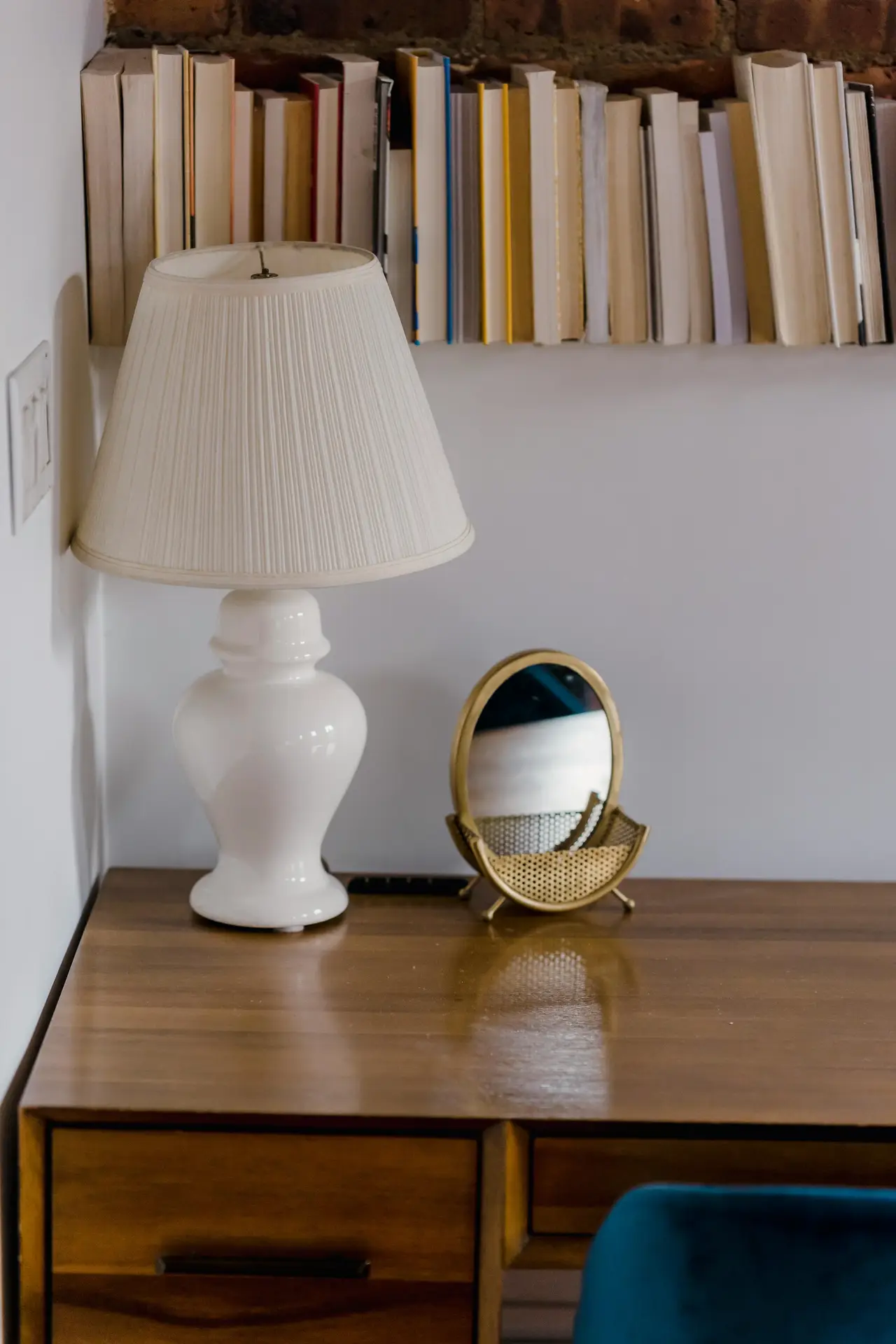 A classic traditional table lamp with a white ceramic urn-style base and a pleated cream lampshade sits on a polished wooden desk. The setting, which includes a small gold vanity mirror and a shelf of books in the background, creates a timeless look that blends well with mid century modern lighting elements.