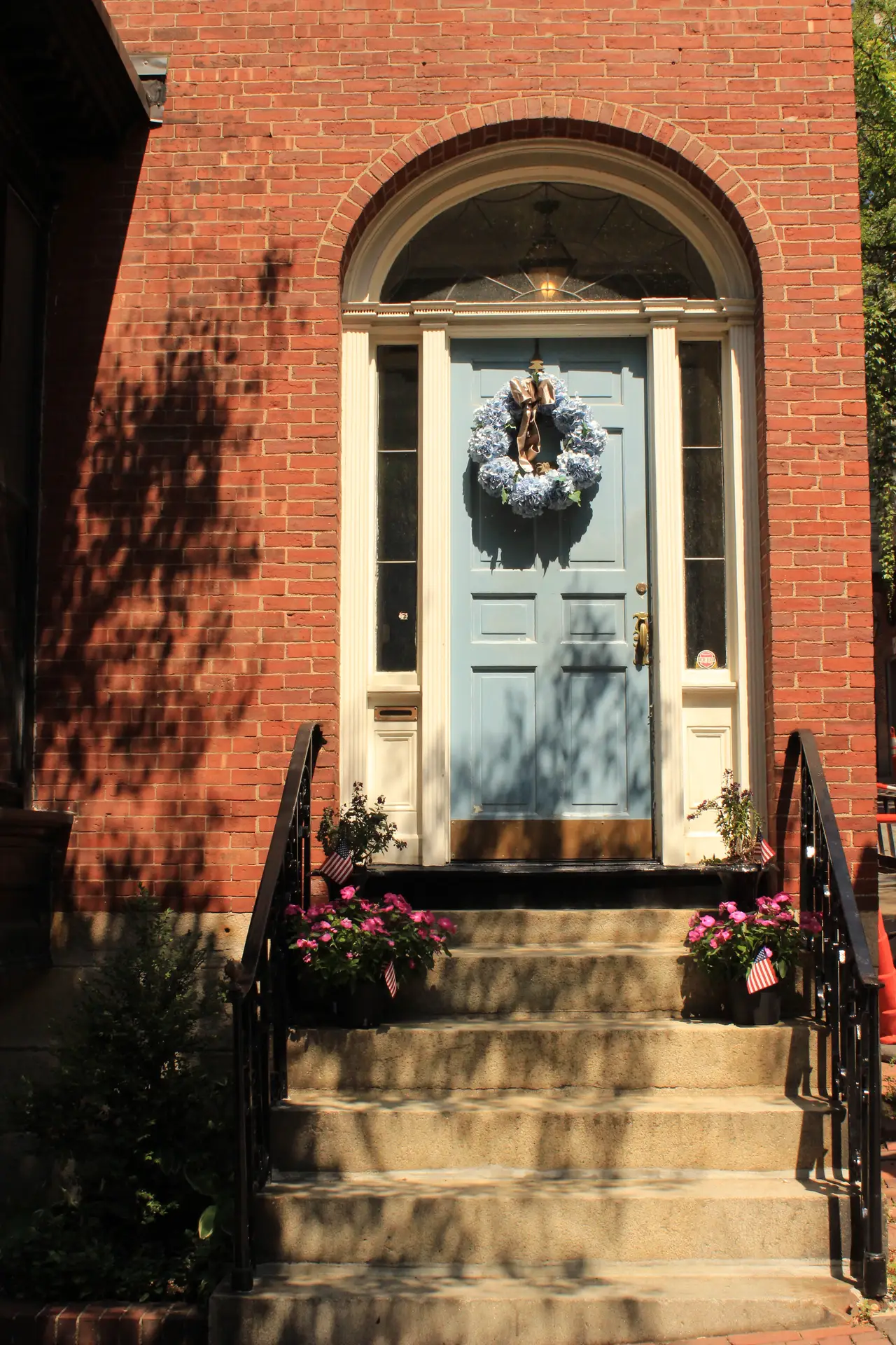 A full blue hydrangea wreath with a large tan bow is centered on a light blue front door, which is framed by a white archway and red brick walls. The sun-drenched entrance features stone steps lined with pink flowers in pots, creating a classic and inviting look for a spring door wreath.
