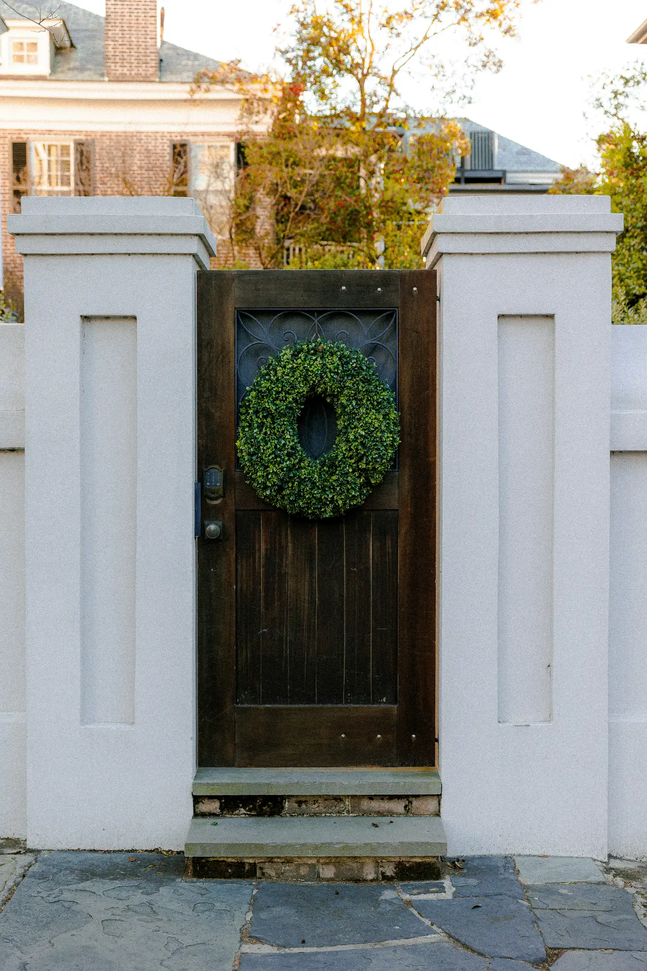 A lush, green boxwood wreath with a dense leafy texture is mounted on a dark wooden garden gate with a decorative metal grate. The gate is framed by two tall white concrete pillars, creating a clean and timeless look for a spring door wreath in a classic outdoor setting.