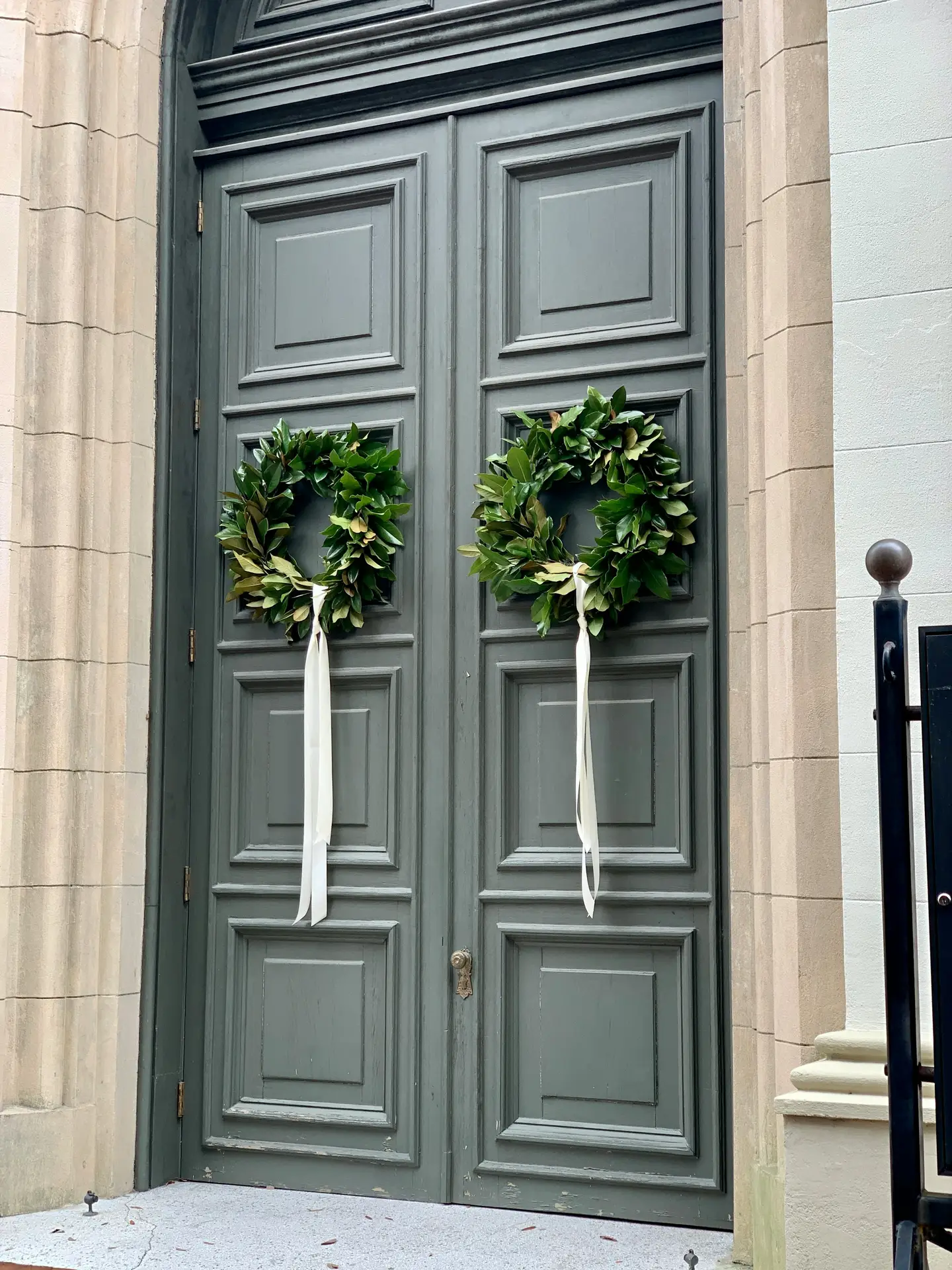 A pair of classic magnolia leaf wreaths with glossy green foliage and long, elegant white ribbons hangs on a grand, grey paneled double door. The symmetrical arrangement provides a timeless and sophisticated look, perfectly illustrating a high-end spring door wreath concept for a stately home entrance.