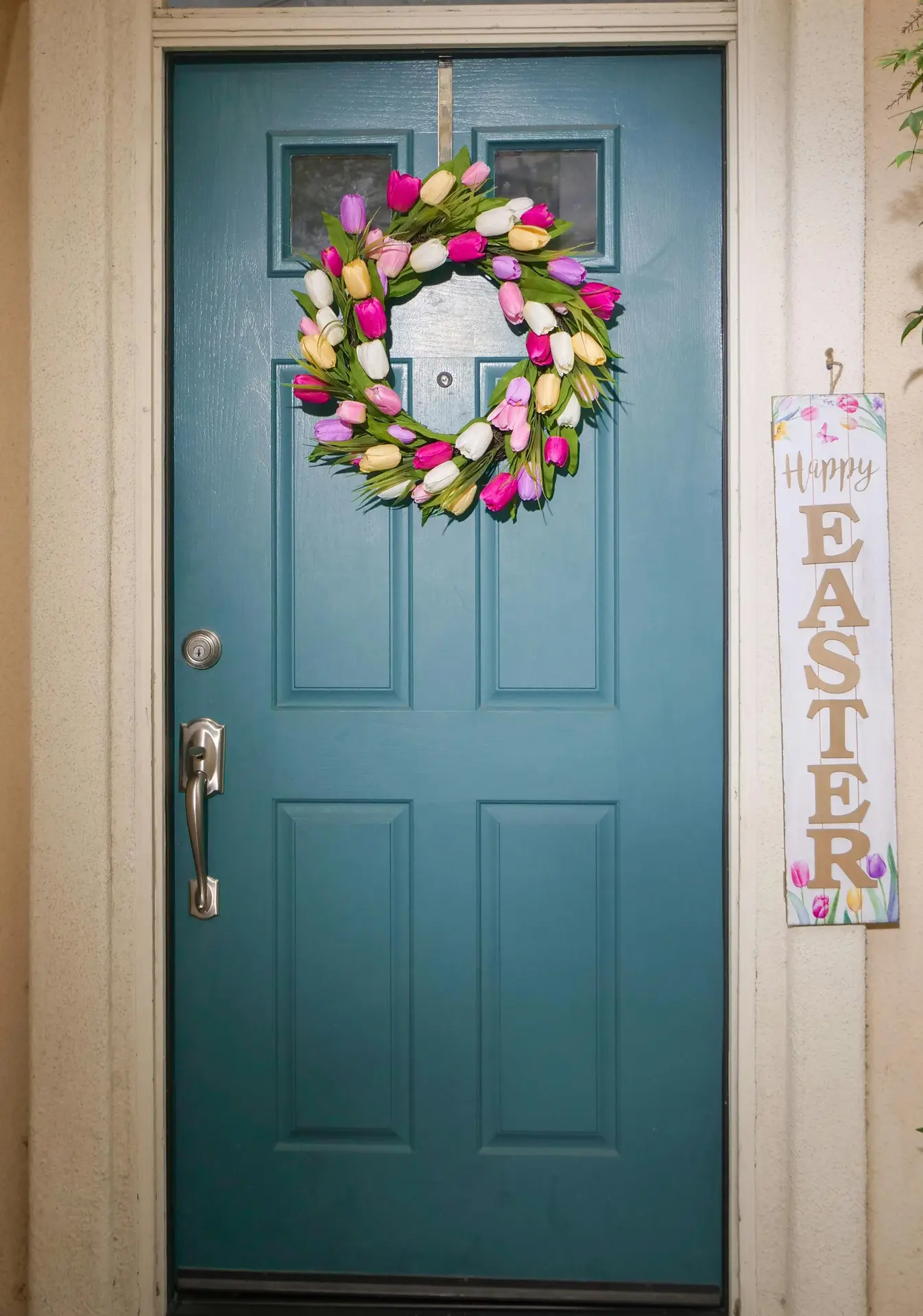 A vibrant colorful tulip wreath featuring pink, yellow, and white tulips with lush green leaves hanging on a teal front door. A decorative 