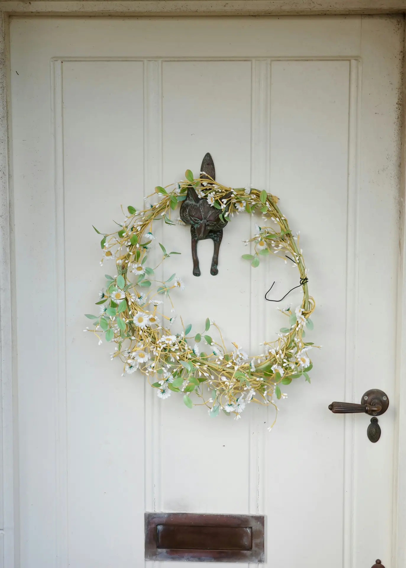 A light and airy daisy hoop wreath made of delicate yellow vines, white daisies, and soft green leaves hangs on an off-white vintage door. The wreath is draped over a unique bronze fox-shaped door knocker, creating a rustic and whimsical look for a spring door wreath.