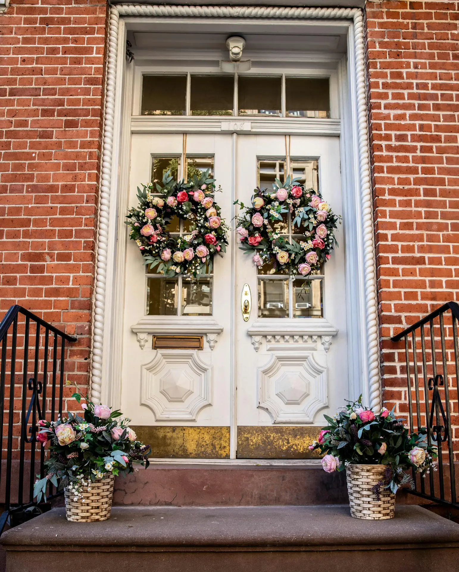 A pair of lush double peony and eucalyptus wreaths featuring soft pink and cream flowers adorn the glass panes of an elegant white double door. Flanked by red brick walls and matching floral baskets on the steps, this symmetrical arrangement serves as a sophisticated example of a spring door wreath for a classic entryway.