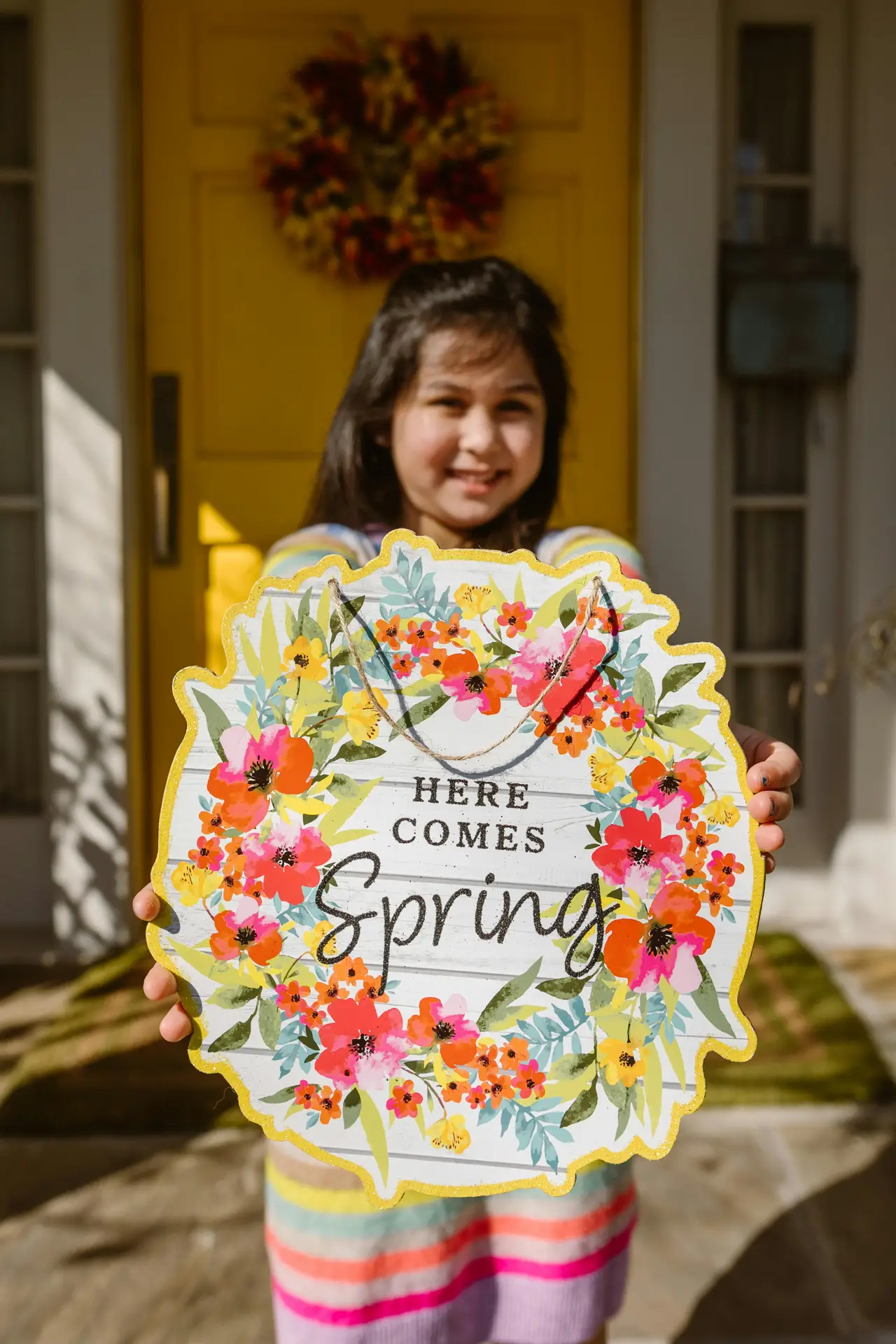 A smiling girl holds a circular floral welcome sign with the text 