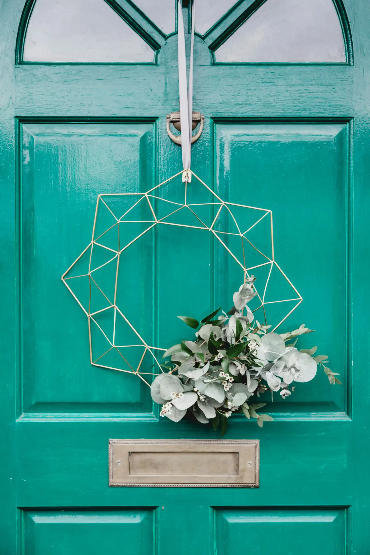 A modern geometric gold wreath with fresh eucalyptus and small white berries is suspended by a light grey ribbon on a vibrant emerald green door. The sharp angles of the metallic frame contrast with the soft, silvery-green leaves, making it a stylish and minimalist choice for a spring door wreath.