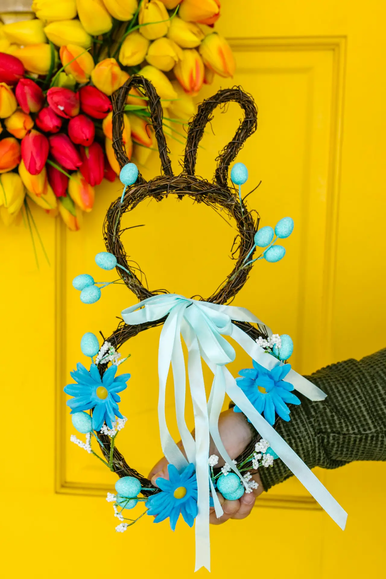A rustic grapevine bunny wreath in the shape of a rabbit silhouette, decorated with bright blue flowers, small white blossoms, and light blue eggs. Held against a vibrant yellow door with a colorful tulip wreath in the background, this playful design offers a creative and cheerful idea for a spring door wreath.