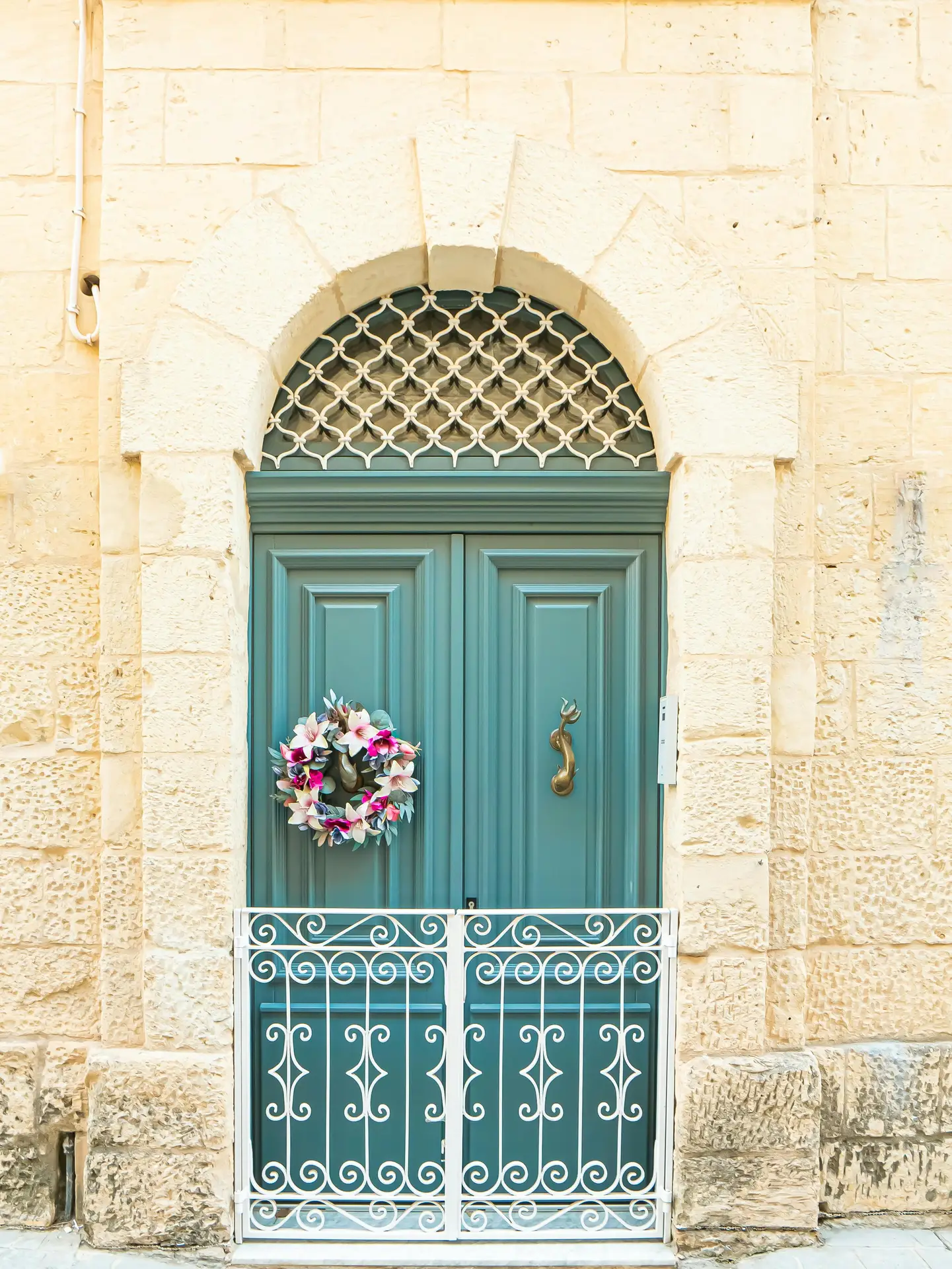 A vibrant lily wreath featuring pink and purple blossoms with soft green foliage is displayed on a teal-colored double door. The door is set within an ancient light-colored stone archway and is partially enclosed by a decorative white wrought-iron gate, making the spring door wreath a beautiful Mediterranean-style accent.