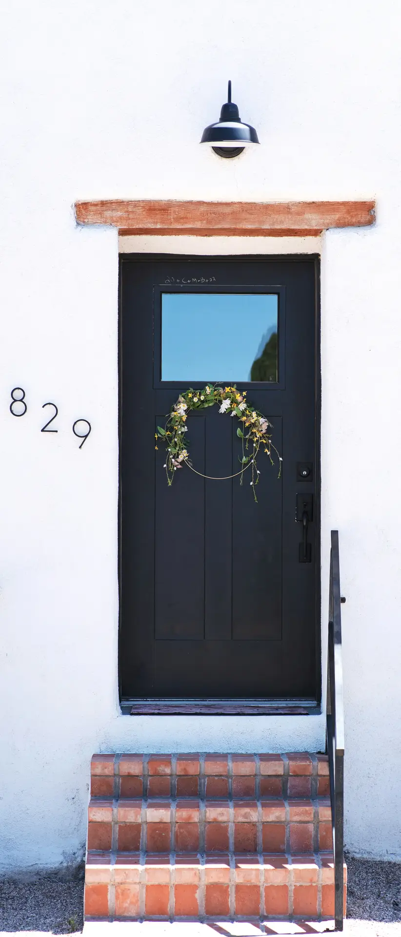 A sleek black front door features a minimalist wildflower hoop wreath with delicate yellow and white blossoms. The thin gold metal ring of the wreath creates a modern, airy look against the dark wood, serving as a perfect example of a contemporary spring door wreath.