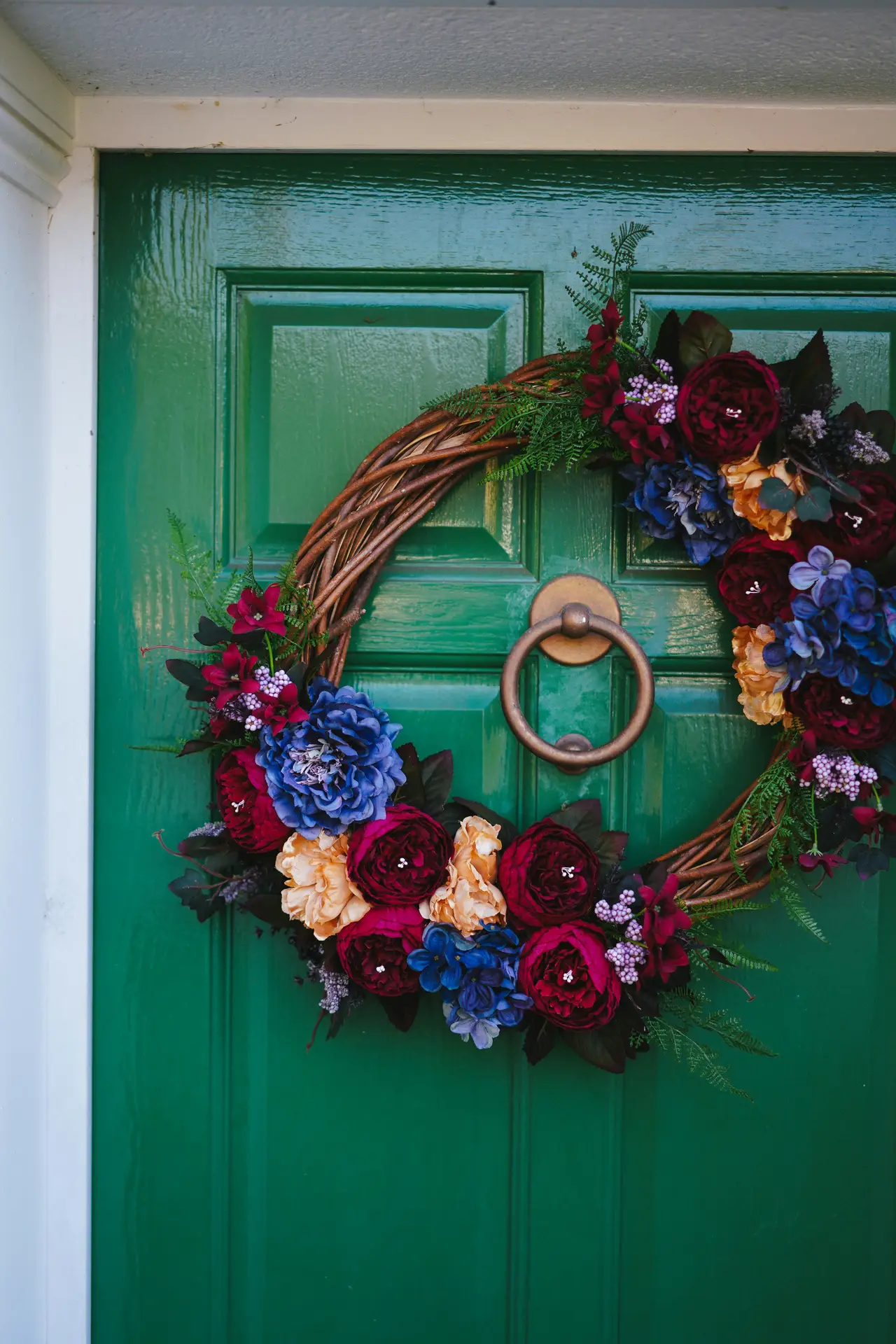 A rustic grapevine base forms a beautiful peony and hydrangea wreath featuring deep burgundy peonies, blue hydrangeas, and soft peach accents. Hanging on a bold emerald green door with a classic brass knocker, this lush floral arrangement offers a moody yet vibrant take on a spring door wreath.