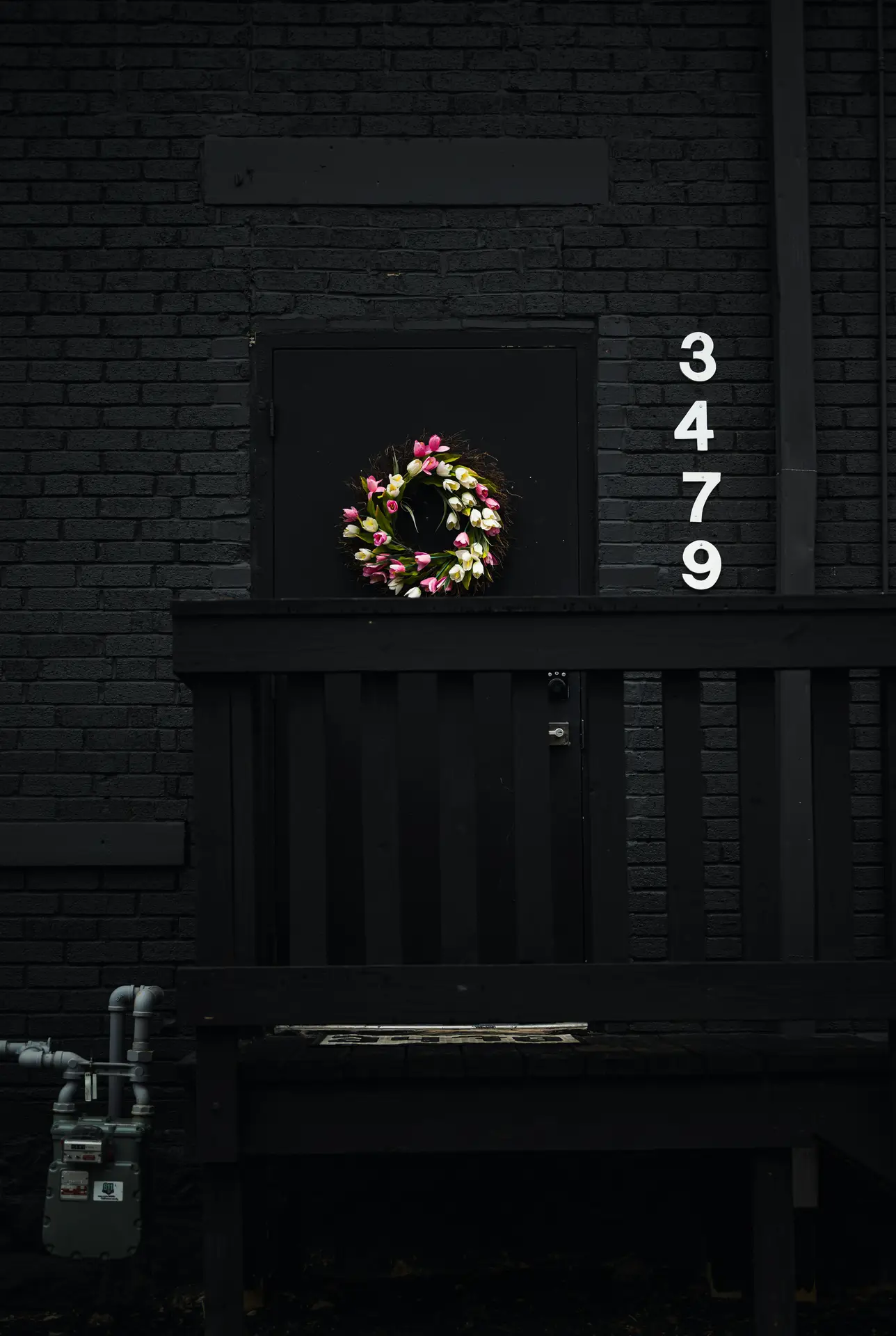 A vibrant pink & white tulip wreath stands out in sharp contrast against a matte black door and dark brick wall. The bright floral arrangement adds a burst of seasonal color to the moody, industrial exterior, serving as a striking and modern spring door wreath.