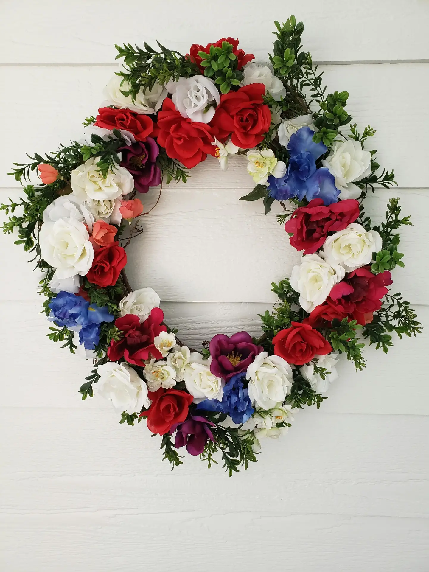 A lush and patriotic red and white roses wreath with blue accents and dark purple blossoms hangs against a white horizontal shiplap wall. The full, circular arrangement is filled with rich green foliage and various floral textures, making it a bold and elegant choice for a spring door wreath.