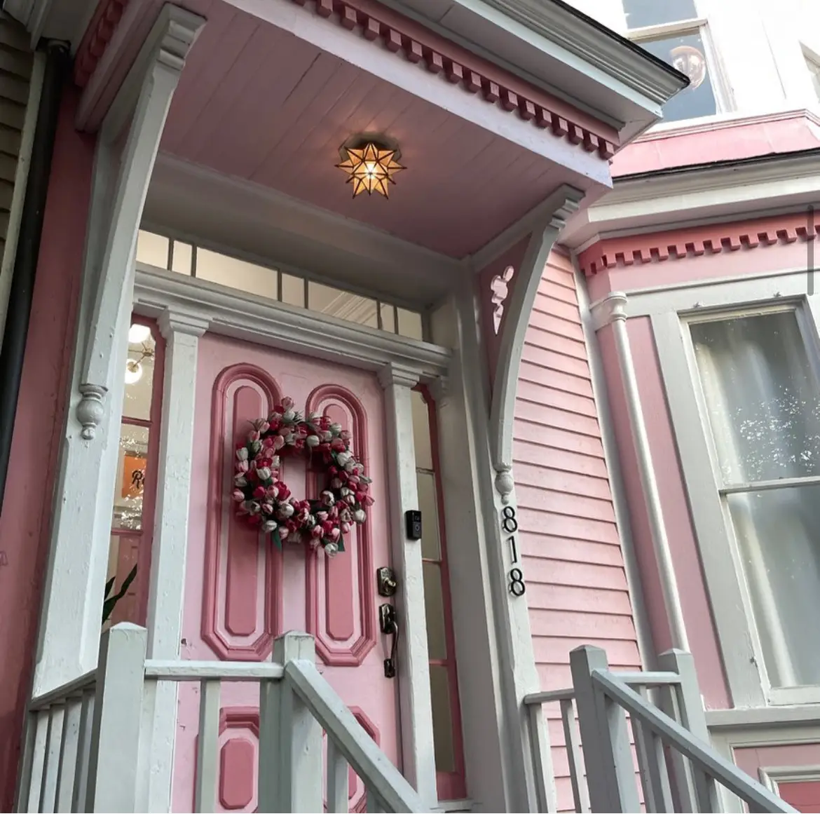 A beautiful vibrant pink tulip wreath hangs on a decorative pastel pink front door, perfectly complementing the charming Victorian-style architecture. This eye-catching spring door wreath adds a welcoming floral touch to the pink wooden siding and white porch details.