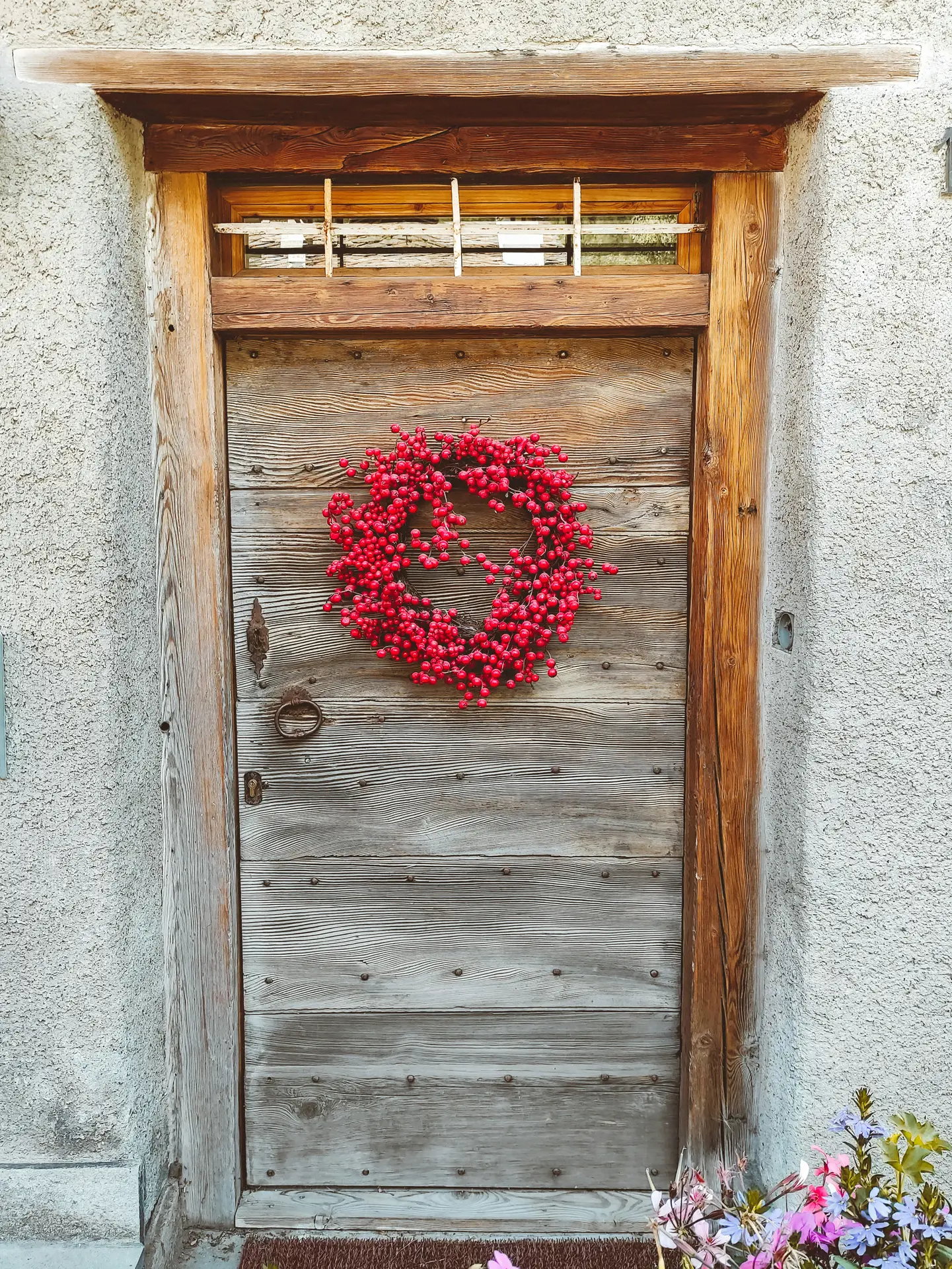 A vibrant red berry wreath stands out beautifully against an aged, rustic wooden door with iron hardware. The dense arrangement of bright berries adds a pop of intense color to the neutral stone wall surroundings, serving as a bold and textured version of a spring door wreath.