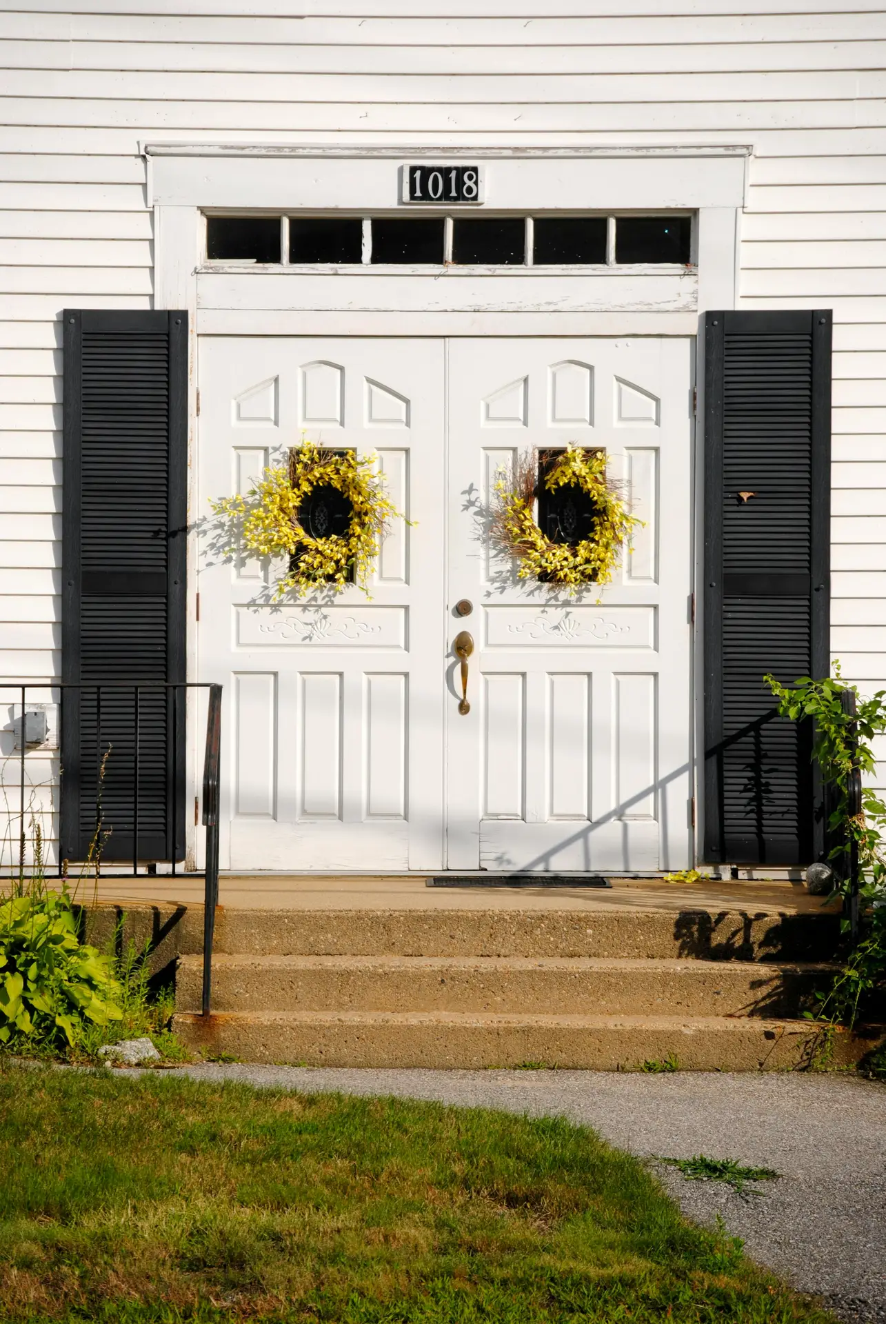 A pair of bright yellow forsythia wreaths adorns the small windows of a classic white double door with black shutters. The vibrant floral arrangements contrast beautifully against the white siding of the house, offering a welcoming and sunny example of a spring door wreath.