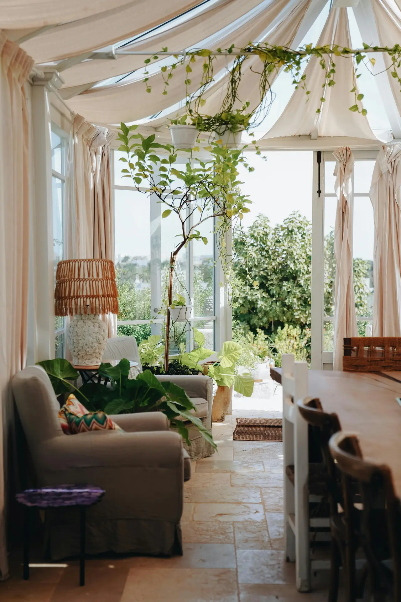 A sun-drenched conservatory filled with inspiration for sun room ideas. The space features a white wooden frame and glass walls looking out to a lush garden. Inside, a daybed is piled high with patterned cushions, throws, and textiles, defining the boho and cozy aesthetic. The room is decorated with numerous hanging plants, string lights, a macramé hanging, lanterns, and a textured kilim rug, creating a warm, jungle-like retreat.