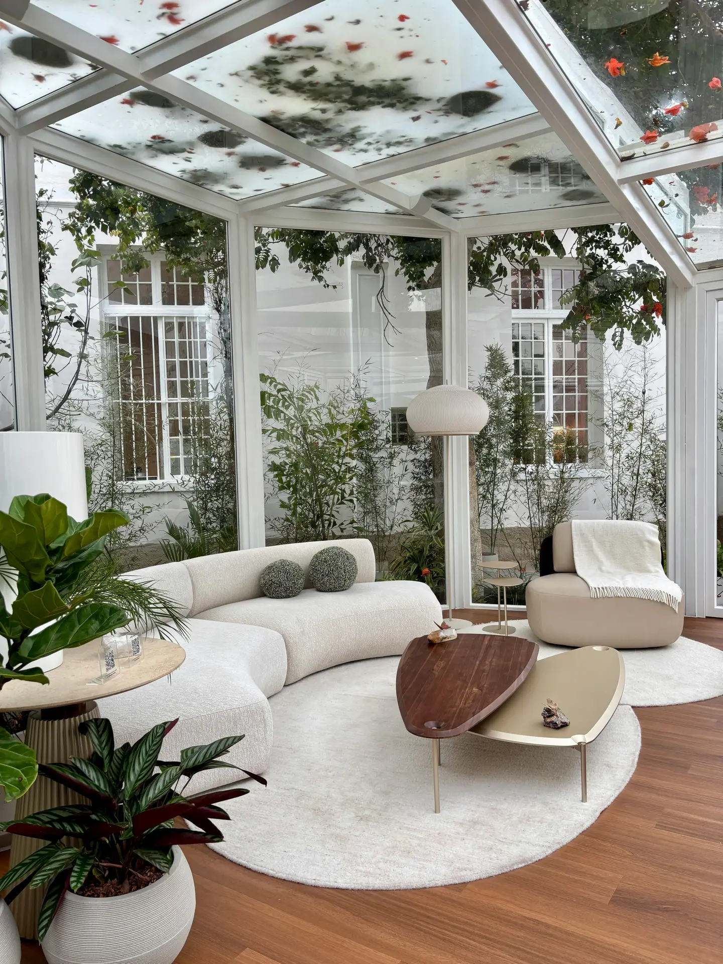 A light-filled space featuring architectural black-framed arched windows that overlook a lush green garden, illustrating modern sun room ideas. Within this airy room, two inviting curvy sofas are paired with an irregular wood coffee table, a jute rug, and various potted plants, creating a comfortable and elegant setting flooded with natural light. The wood beams and white walls complete the serene atmosphere.