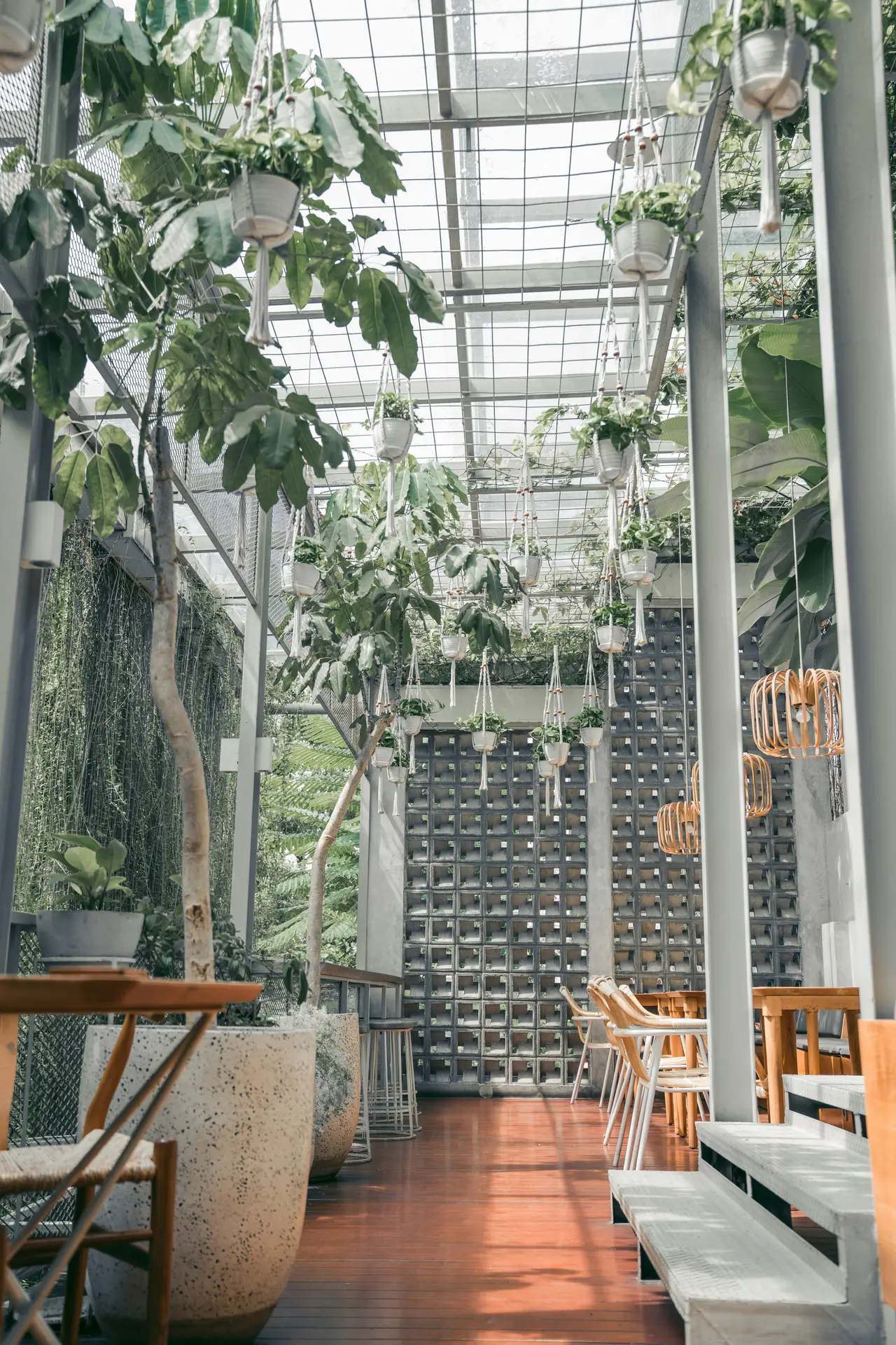 A light-filled, cozy sunroom corner is presented as a prime example of sun room ideas. Built with natural wood posts and beams, the glass walls and roof allow natural light to fill the space. Numerous hanging plants on the ceiling are suspended from the beams. Below, a wicker armchair with cream-colored cushions is nestled among a large Monstera deliciosa, assorted ferns, and smaller potted plants on shelves. The view through the glass is of a vibrant, verdant garden.