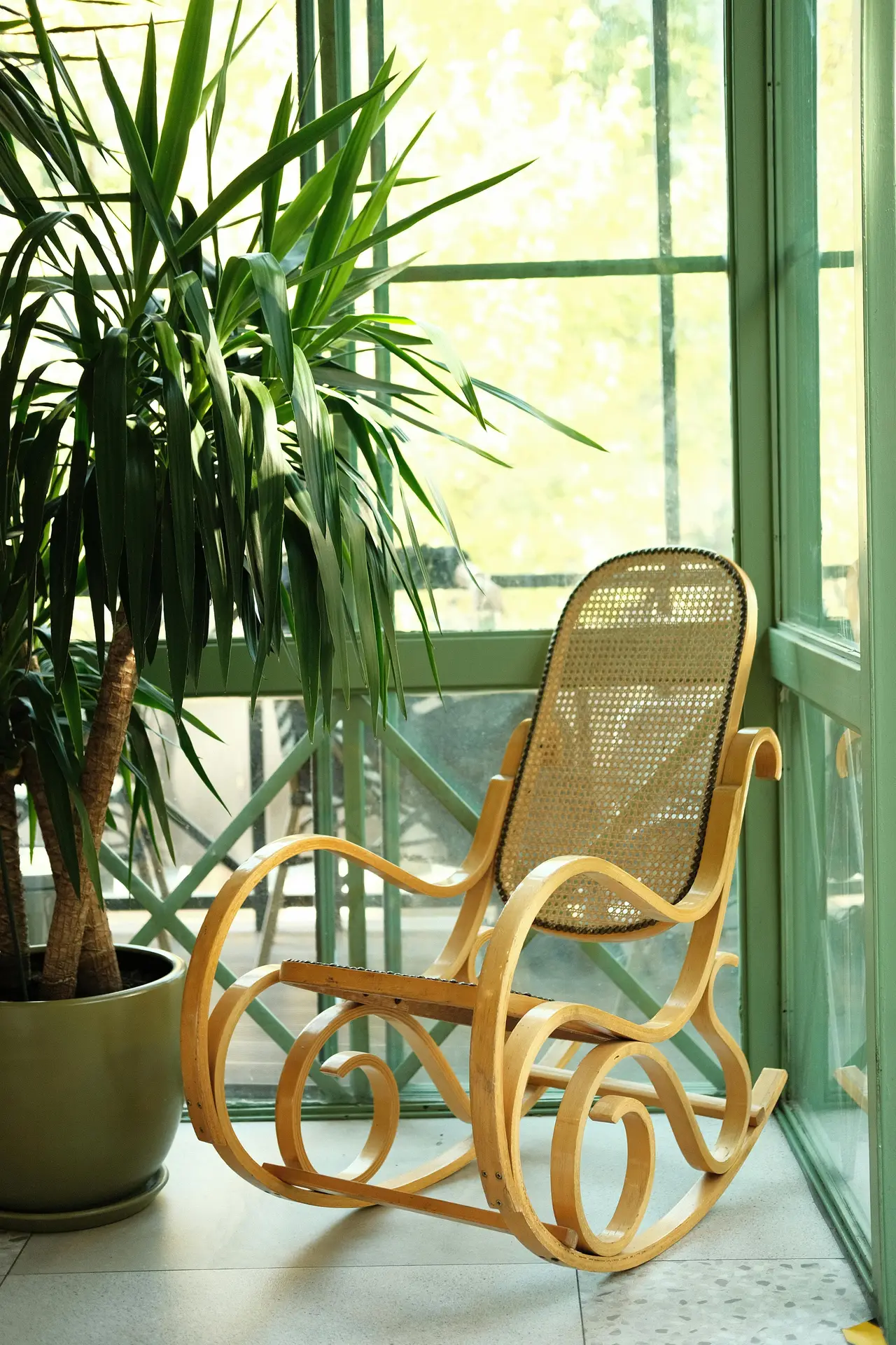 A peaceful, light-filled sunroom corner featuring a vintage, light blue wooden rocking chair draped with a thick, cream-colored natural wool throw and a small patterned lumbar pillow. A pair of worn leather slippers rests on the natural flagstone floor in front. Large white-framed glass windows and glass roof panels provide a view of a lush green outdoor garden, with sunlight creating bright spots. Next to the chair is a rustic tree-slice side table with a ceramic mug and a closed book. Thriving potted plants, including a large snake plant, are nearby. This scene illustrates inviting sun room ideas for creating a cozy spot and how to comfortably integrate a rocking chair for relaxation.