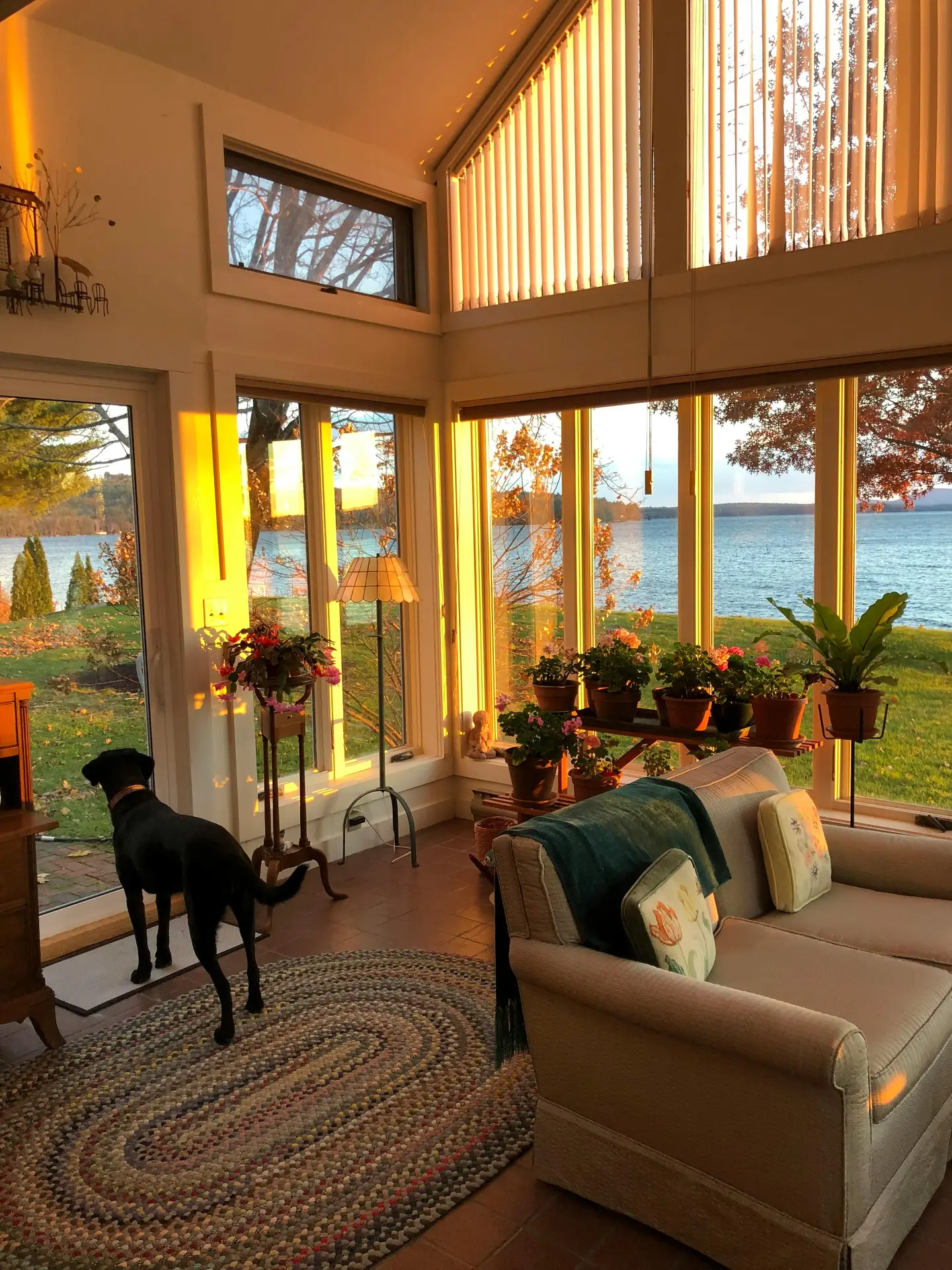 A view from within a cozy sunroom featuring a glass roof and wall panels supported by massive, natural log beams and posts. The interior is furnished with a large, carved wood-frame sofa covered in neutral-toned cushions and a textured, multi-colored striped blanket, resting on a stone-tile floor. A round wooden side table holds a lamp with a woven base and a black vase, with baskets stacked nearby. Large glass windows provide a stunning panoramic vista of a wide lake and a dense forest shoreline on the opposite bank under a bright sky, illustrating inviting sun room ideas with authentic lake house and cabin vibes.