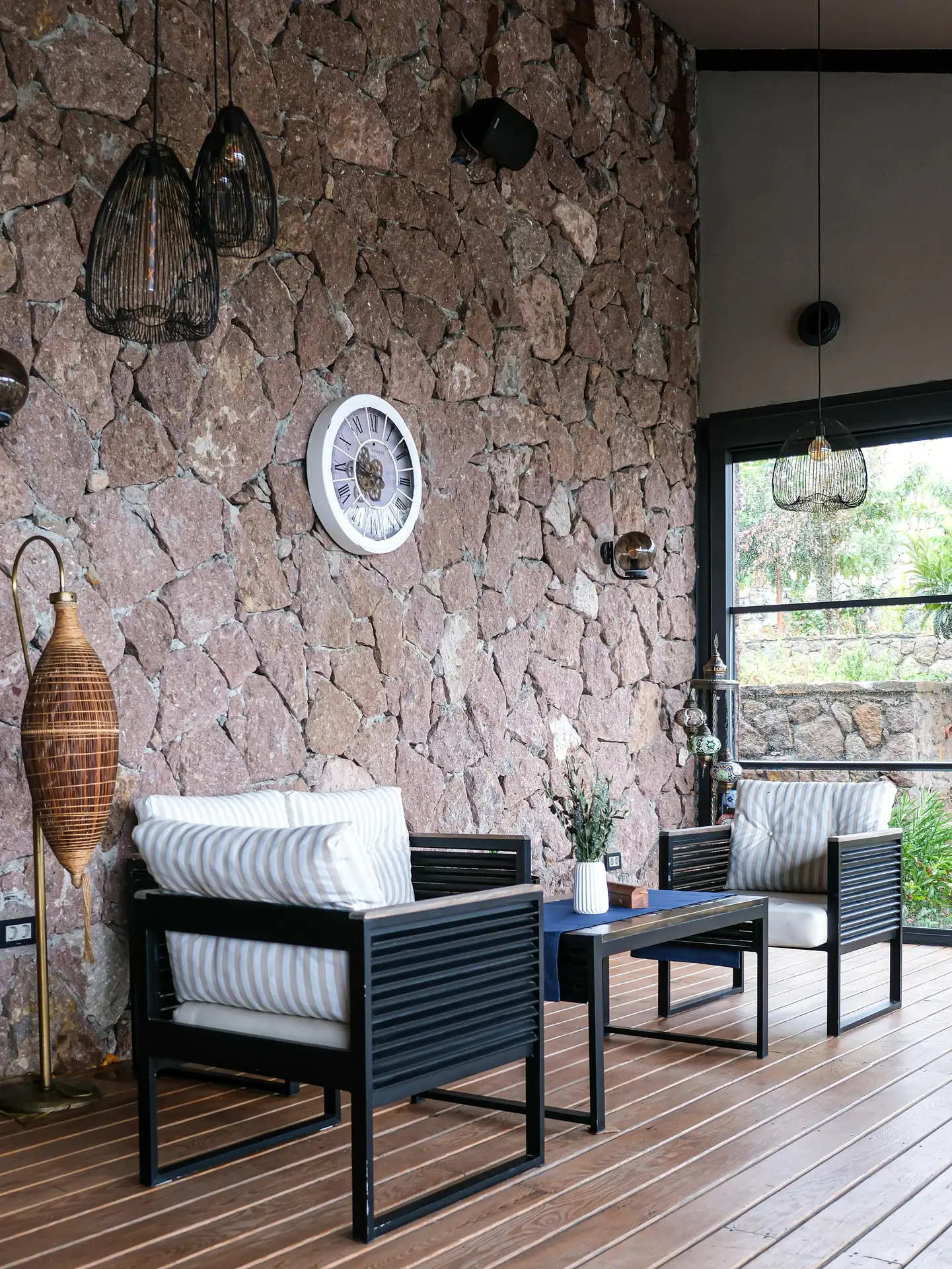 A sun-drenched conservatory, offering inspiration for sun room ideas, features full-height black-framed glass walls and a glass ceiling supported by a network of exposed natural wood beams. The modern and industrial style interior includes a grey modular sofa with simple cushions, two round wooden coffee tables, a black wire lamp, and a striped jute rug on a concrete floor. Multiple potted plants, including a large Monstera, add life, while the outdoor view of green trees is visible. Sunlight is abundant.