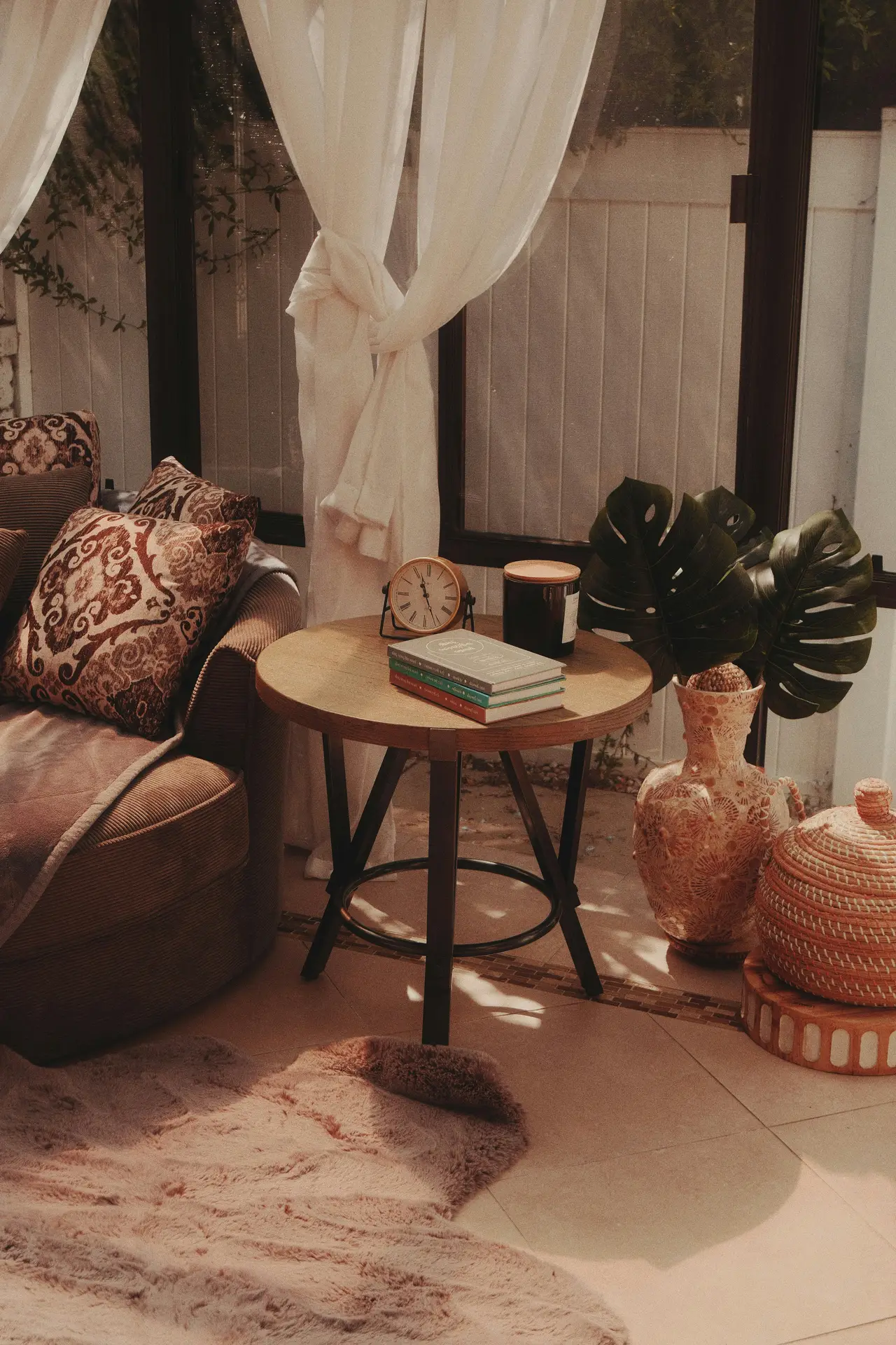 A cozy, sun-drenched reading nook is perfectly integrated into a light-filled sunroom, offering excellent sun room ideas for relaxation. Large, framed glass windows with expansive views of a lush green garden define the space, allowing copious natural light to pour in. The comfortable armchair is deeply upholstered in white fabric and abundantly layered with a cream-knit blanket, a rust-orange throw, and a vintage striped textile. A patterned round cushion rests on the seat. Next to the chair, a rustic wood side table holds a stack of books, a pair of eyeglasses on top, and a steaming ceramic mug. A large potted Monstera plant stands in the background. A macrame wall decoration hangs to the left, and a patterned rug covers the floor with comfortable slippers nearby.