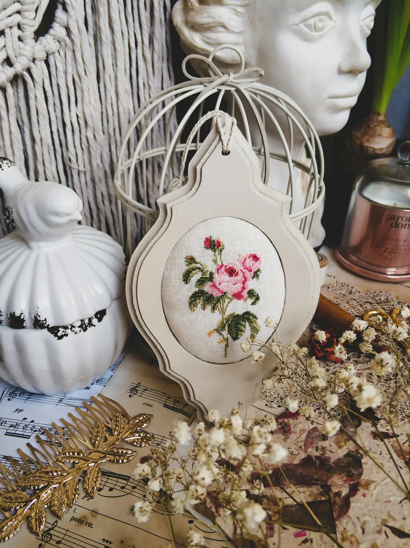 An artistic arrangement of vintage garden decor features various ceramic elements and embroidered images. In the center is a delicate cross-stitch embroidery of pink roses in a cream-colored frame, surrounded by a white ceramic bird jar, a bust of a woman, a wire birdcage, and golden accessories laid out over vintage sheet music and dried flowers.