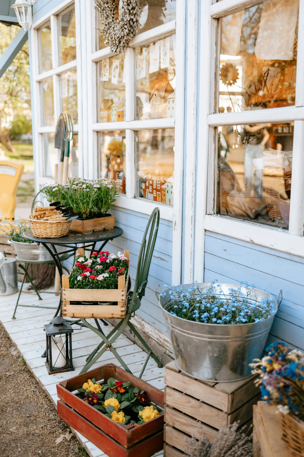 A detailed view of a vertical garden arrangement on an aged, white-painted wooden wall, demonstrating vintage garden decor. The display includes dried flowers in a hanging metal basket and a distressed galvanized bucket below. In the middle, a mounted wooden crate is repurposed as a planter, holding assorted green leafy plants and a single pink bloom. The scene is full of texture, showcasing flowers in repurposed crates and buckets in a shabby chic style.