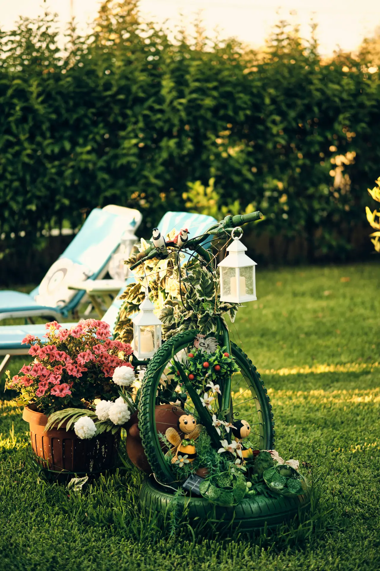 A vibrant photograph capturing a creative piece of vintage garden decor set on a green lawn with a blurred background. A central, functional green bicycle garden decor piece is adorned with multiple items. It features several white metal lanterns with glass panels hanging from the handlebars and frame, small bird and bee figurines, and is decorated with artificial ivy and white flowers. Potted plants are integrated: a large terracotta pot with dense pink and white flowers sits next to the rear wheel, while a smaller plant in a green tire planter is at the base. Blue lawn chairs and a hedge are visible in the distance under daylight.