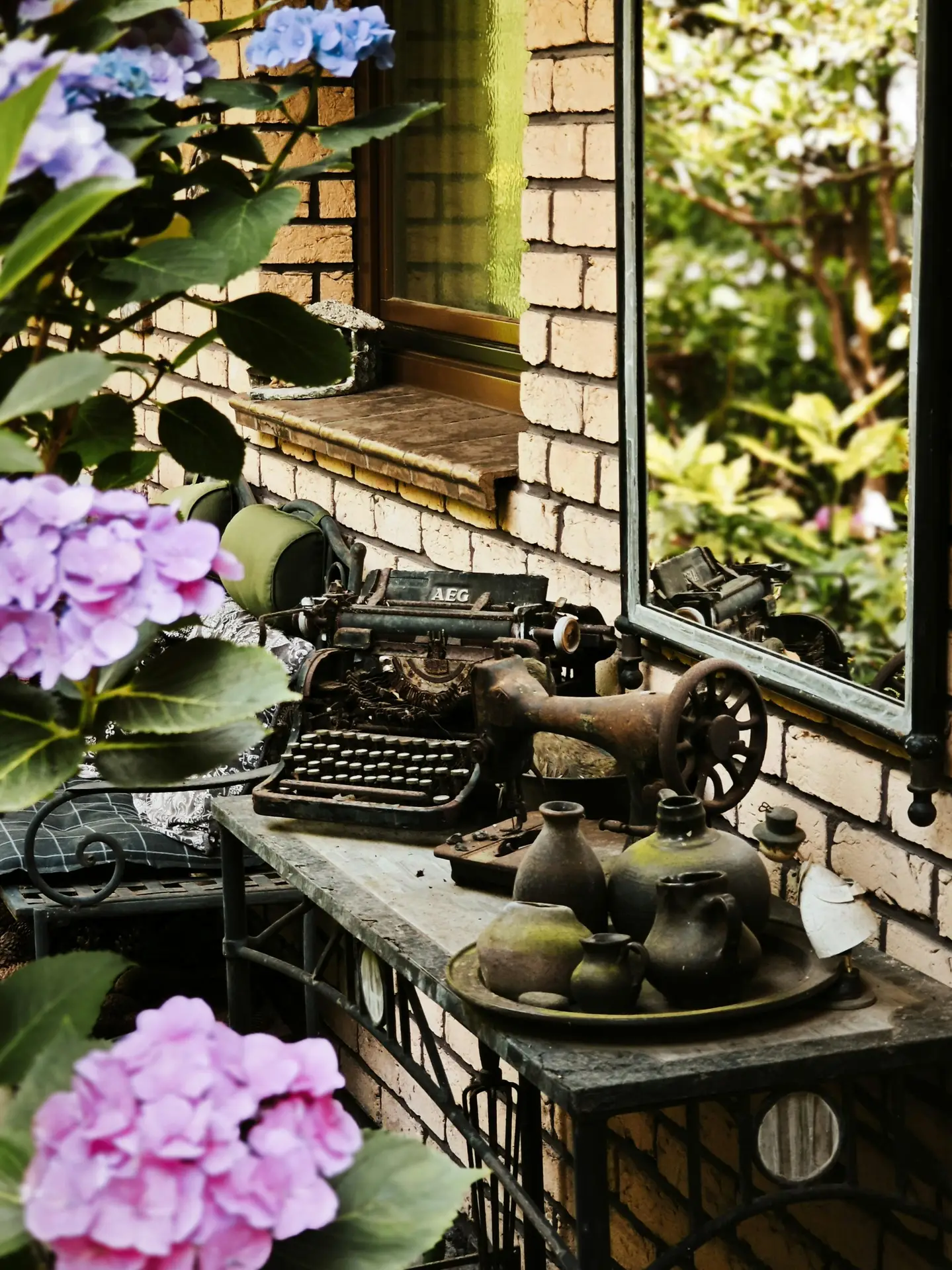 A charming scene of vintage garden decor arranged on a dark metal table against a textured brick wall and a large window. The display features an antique AEG typewriter and a rusty, manual old sewing machine. Adjacent to them is a tray with an assortment of weathered black ceramic pitchers and bowls. In the foreground and background, lush blue and purple hydrangeas with green leaves add vibrant color. A large mirror on the right reflects the scene and more blurred greenery, enhancing the antique and natural atmosphere.