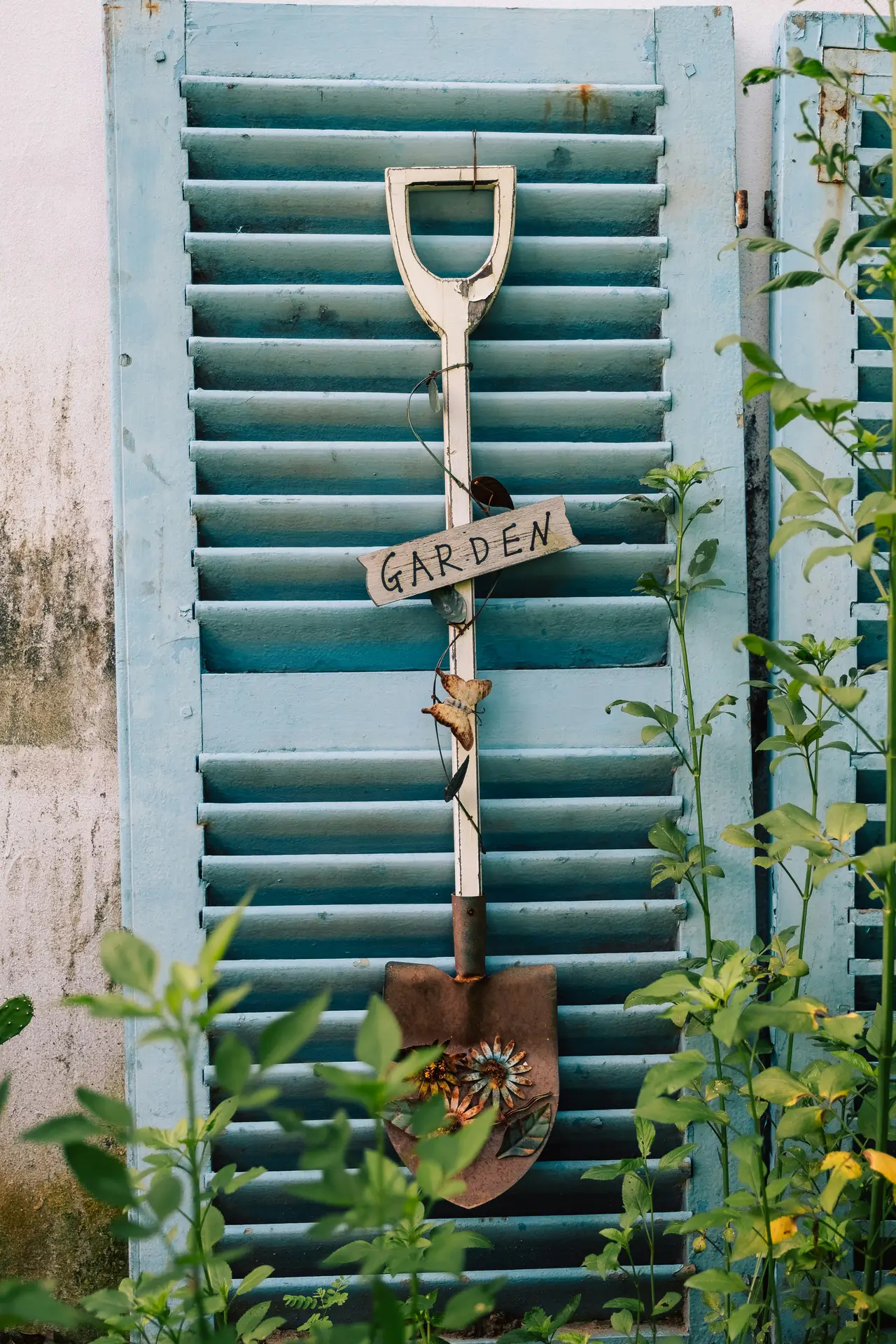 A piece of unique vintage garden decor shows a vertically suspended old shovel with a sign hanging on its white wooden handle. Below the rustic wooden sign reading 'GARDEN' and some butterfly charms, the shovel features a heavily rusted metal blade adorned with decorative welded flowers. It is set against distressed, light-blue louvered shutters and partially obscured by green garden plants.