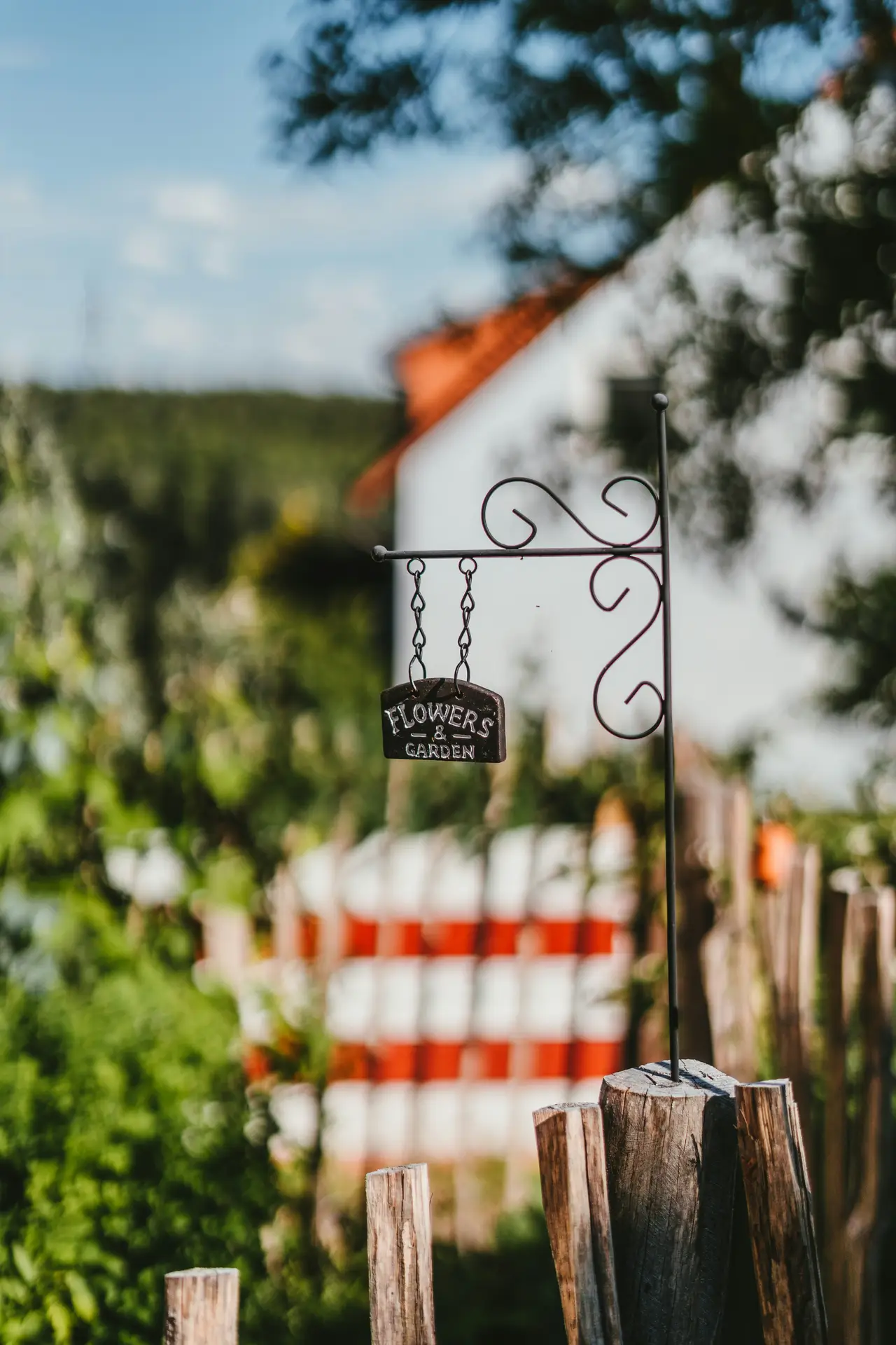 A close-up photograph of vintage garden decor featuring a small, dark metal ornate sign with nostalgic charm. It is inscribed with the white text 'FLOWERS & GARDEN' and hangs on chains from a black iron bracket. The sign is attached to a rustic wooden fence post, with a blurred garden, greenery, and a red-and-white-striped background elements visible in the background.