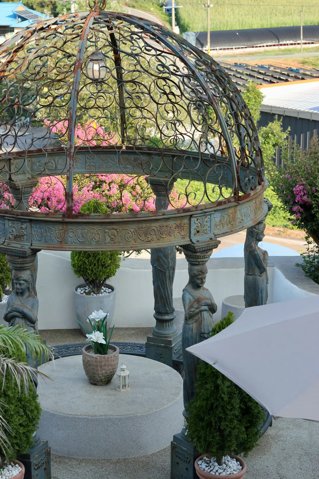 A rustic, wooden-framed vintage garden pavillon with weathered blue painted posts and a solid roof stands at the end of a stone path in a lush garden. Two white-painted wooden benches with striped cushions face each other inside the pavilion, which is adorned with potted plants, a hanging lantern, and textiles, epitomizing a cozy and welcoming vintage garden decor theme.