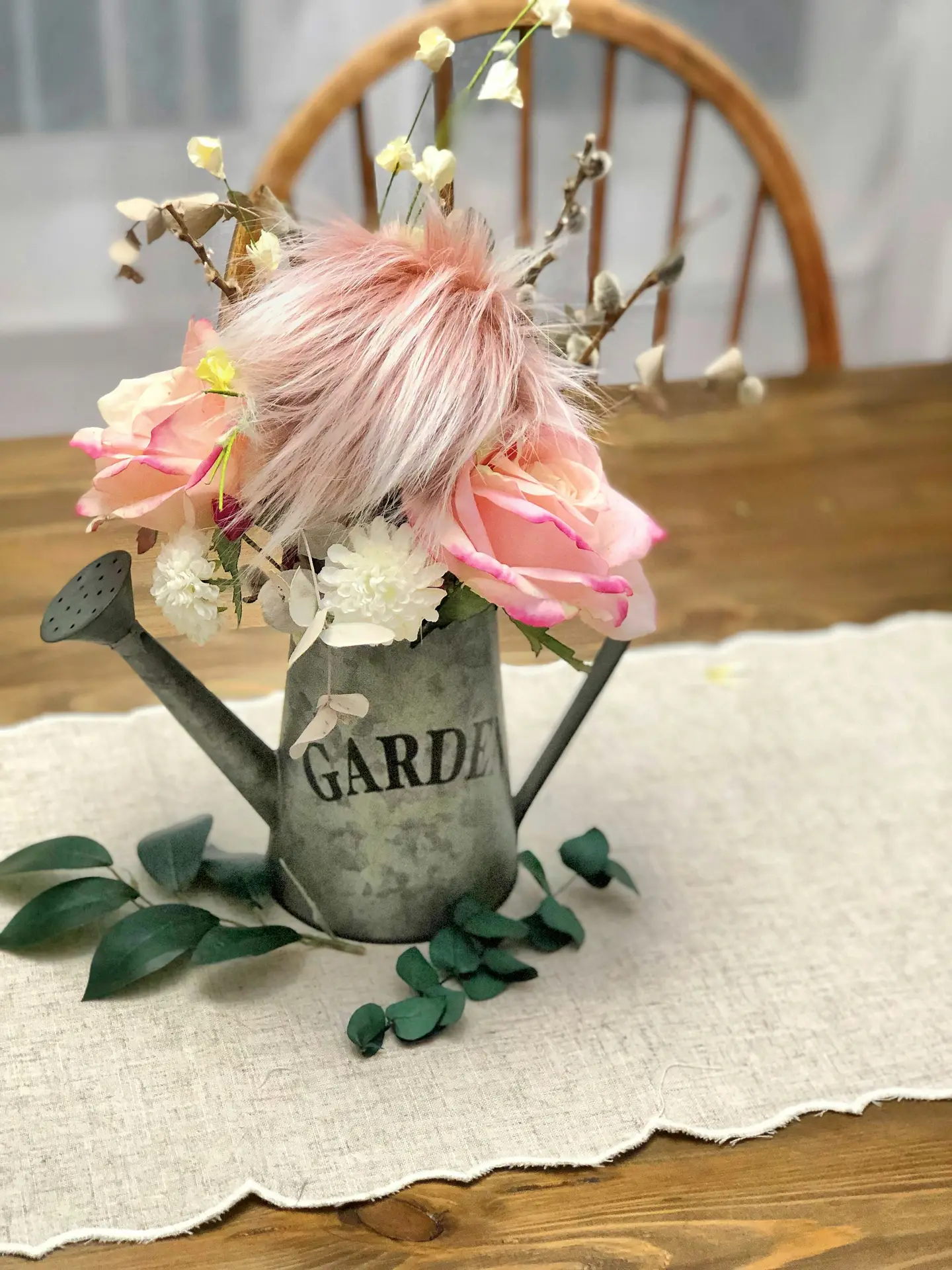 A decorative indoor arrangement of vintage garden decor features a small, weathered zinc watering can as a flower vase. The watering can, embossed with the word 'GARDEN', is filled with large pink roses, small white blossoms, pussy willow branches, and a fluffy pink pom-pom. It sits on a linen table runner over a wooden table, with a few green eucalyptus-style leaves scattered at the base and a wooden chair in the blurred background.