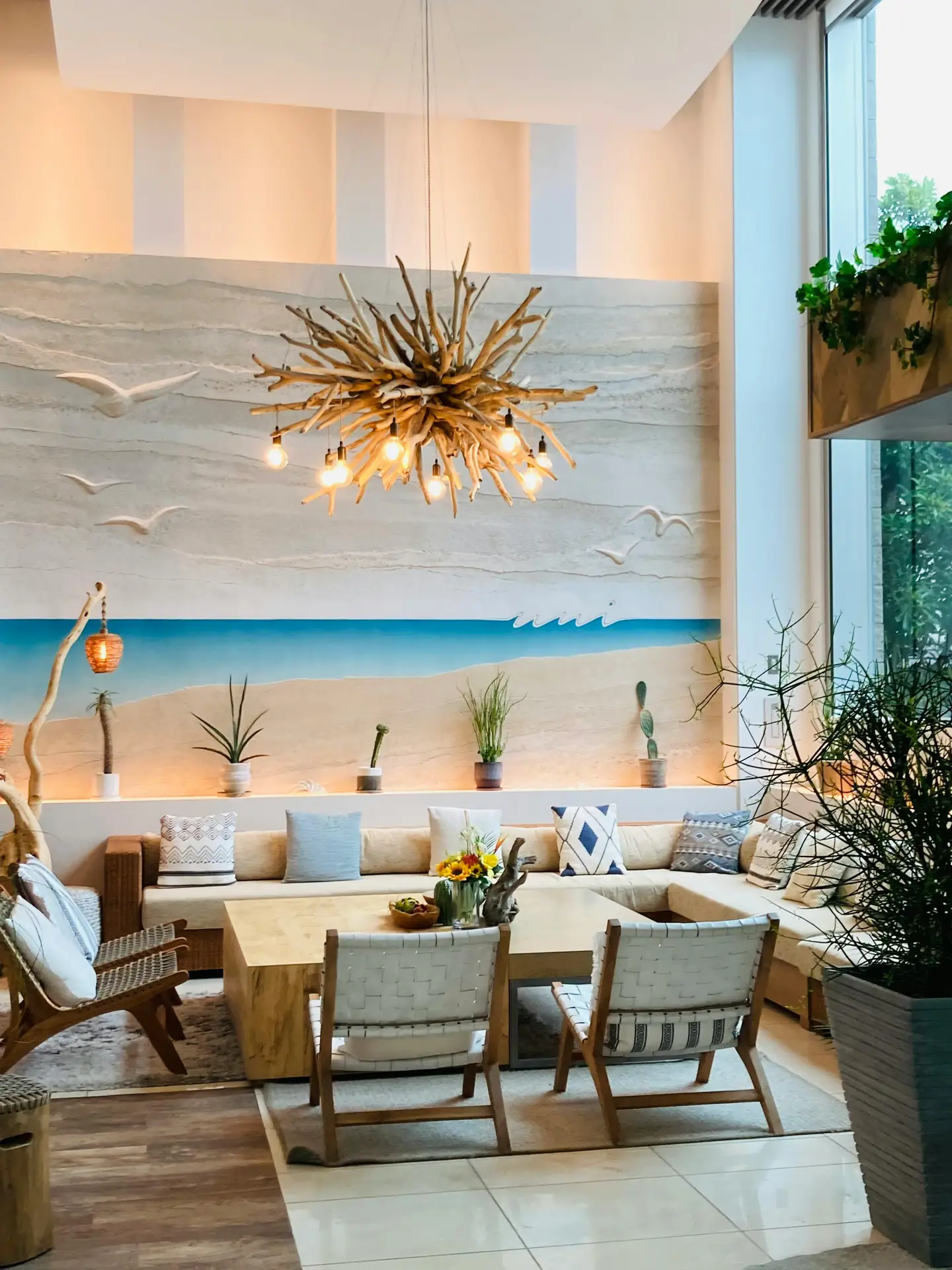 A spacious and airy interior showcasing coastal living room ideas that radiate relaxed beach vibes. The room features a large sectional sofa with light blue and patterned pillows, centered around a heavy wooden coffee table. A massive driftwood chandelier with exposed bulbs hangs from the high ceiling, serving as a striking focal point. The back wall is decorated with a large beach-themed mural showing sand, blue water, and white seagulls in relief, while a ledge below holds several small potted plants and cacti. Woven chairs and natural textures complete this bright, ocean-inspired sanctuary.