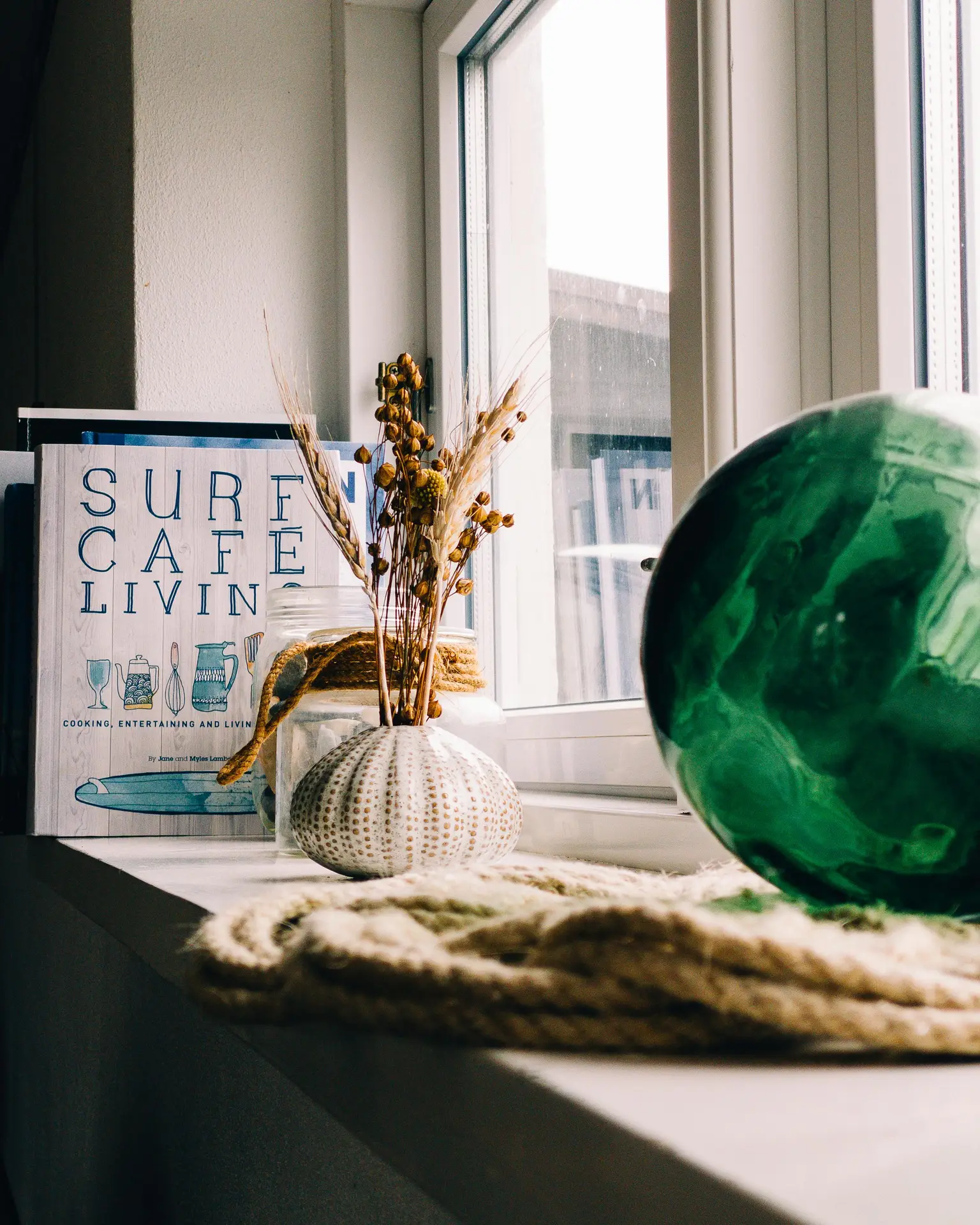 A bright and airy space featuring coastal living room ideas with a focus on a distressed white sideboard. This piece of furniture holds various coastal decor details, including a glass lamp base, a framed line drawing of sea oats, and a white, wave-patterned tray filled with starfish, white coral, and smooth grey and white stones. In the background, a white slipcovered armchair sits near a window, flanked by a tall snake plant. Natural daylight illuminates the soft, natural tones and textures of the jute rug and the sand-colored walls.