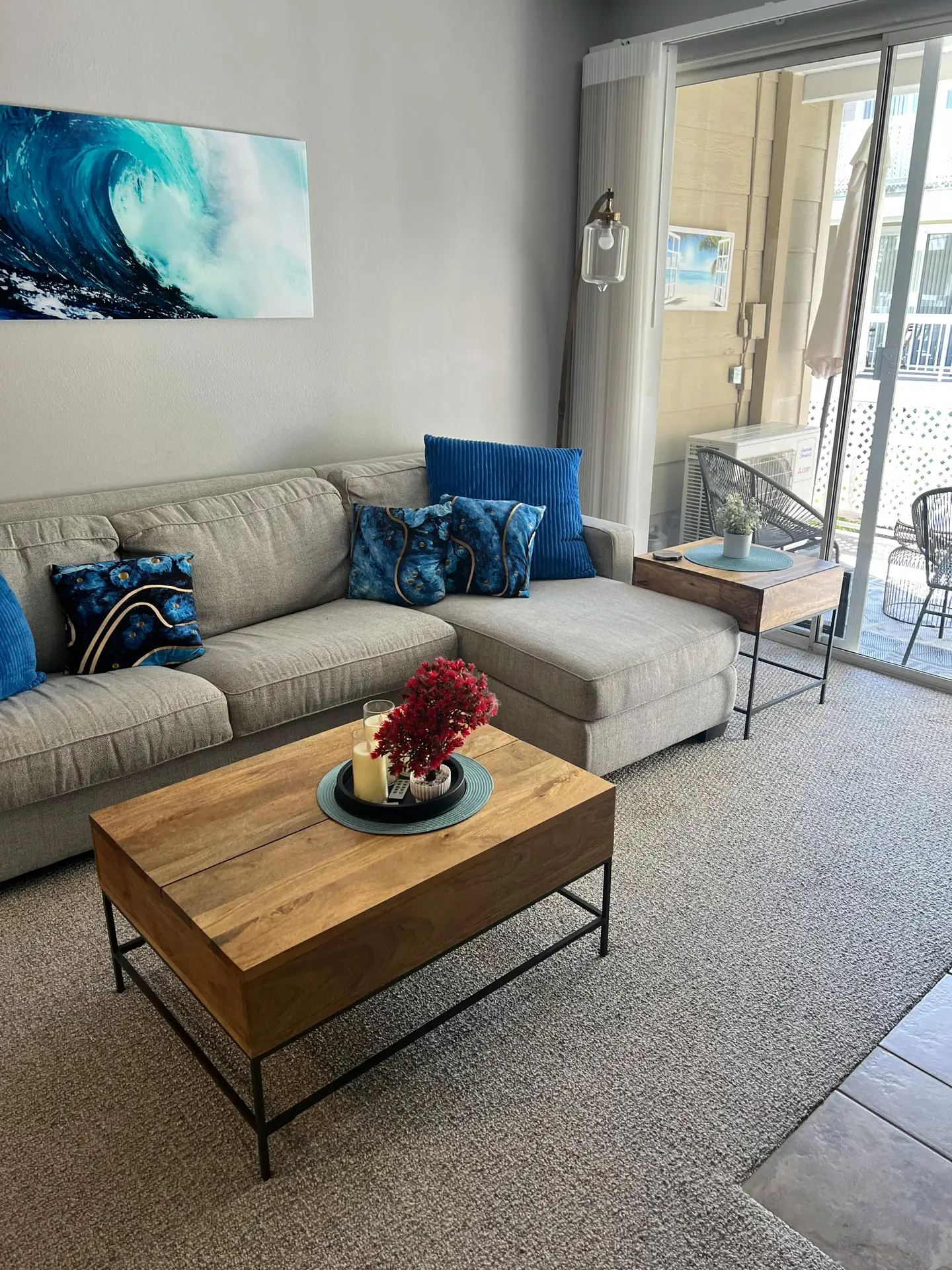A modern living space incorporating coastal living room ideas with a refined Driftwood Grey & Blue color palette. A comfortable light grey sectional sofa is styled with various blue accent pillows featuring wave-like patterns and deep navy textures. In the center, a solid wooden coffee table with a black metal base holds decorative candles and a vibrant red coral-like ornament. The nautical theme is further emphasized by a large canvas art piece depicting a crashing ocean wave on the wall, while sliding glass doors open to a balcony, filling the room with natural light.