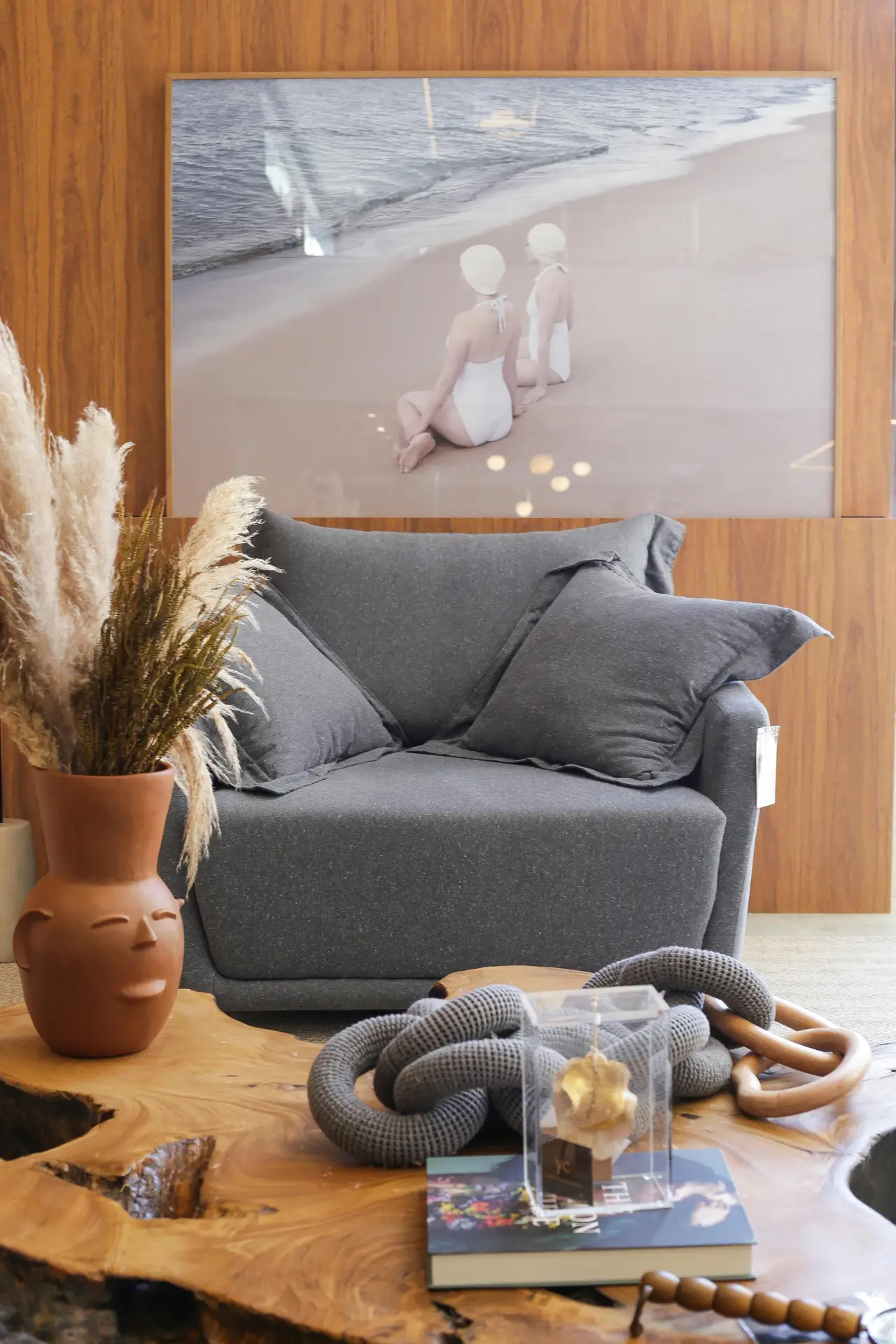 A warm and textured close-up of a living space vignette illustrating coastal living room ideas. The foreground features a rich combination of driftwood grey and wood elements, starting with a thick, rough-cut reclaimed wood console table. On the table, a unique round mirror with a frame made of interwoven driftwood grey branches reflects the soft light, accompanied by a small pile of round, polished river stones and a green glass bottle with dried stems. Next to the table sits a large, textured terracotta pot with a snake plant, and in the blurred background, a cozy armchair is visible against a neutral-colored wall, all contributing to a weathered, natural aesthetic.