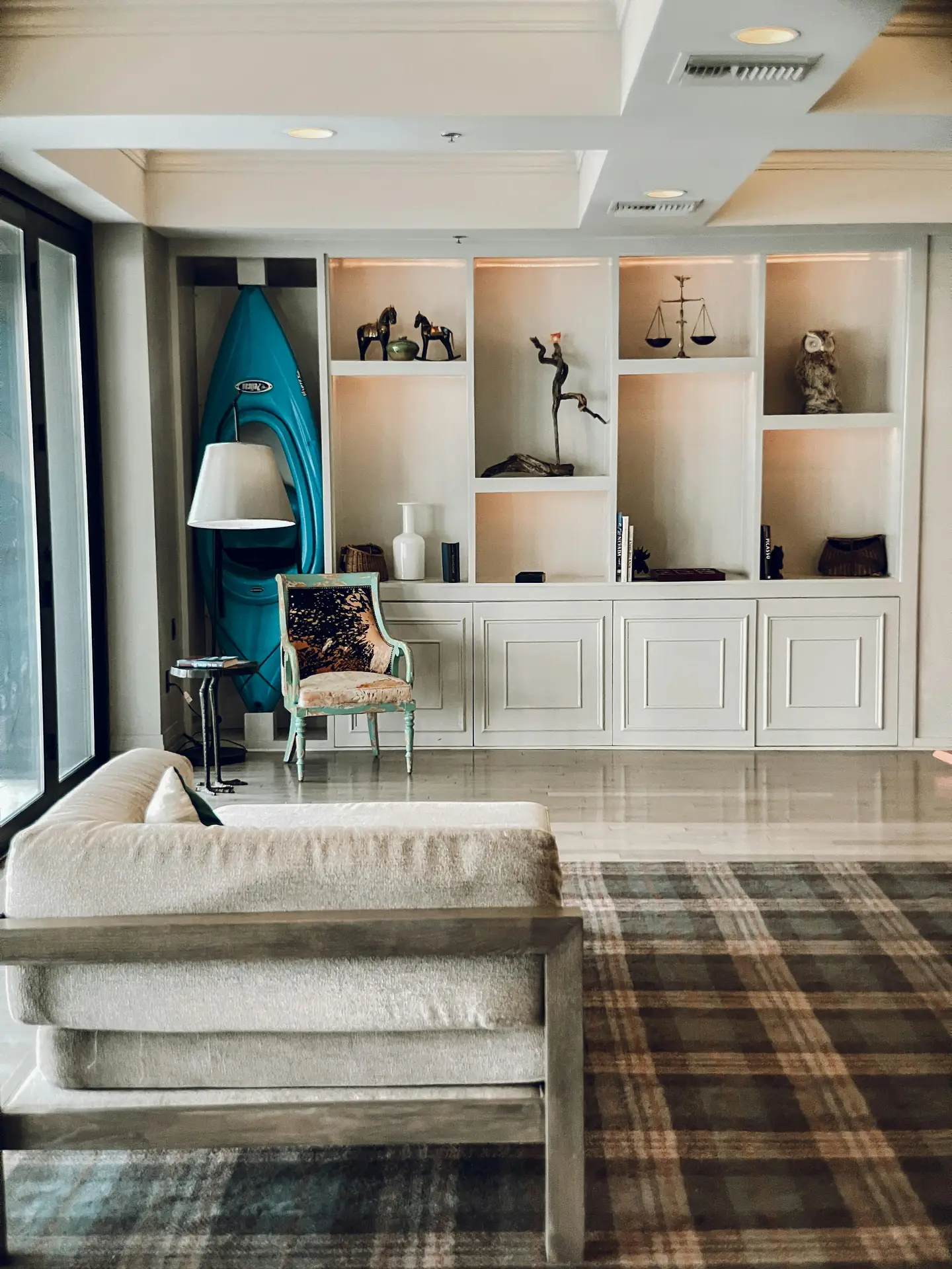 A vibrant close-up featuring charming and quirky funny coastal details, part of inspiring coastal living room ideas. Clustered on a rustic wooden shelf are various character-filled objects: a framed vintage print of three mermaids, a distressed wooden whale figurine, and a pair of playful, round ceramic sculptures resembling stylized puffer fish. Woven baskets are stacked below, all set against a white-painted brick wall, creating a playful, laid-back seaside atmosphere.