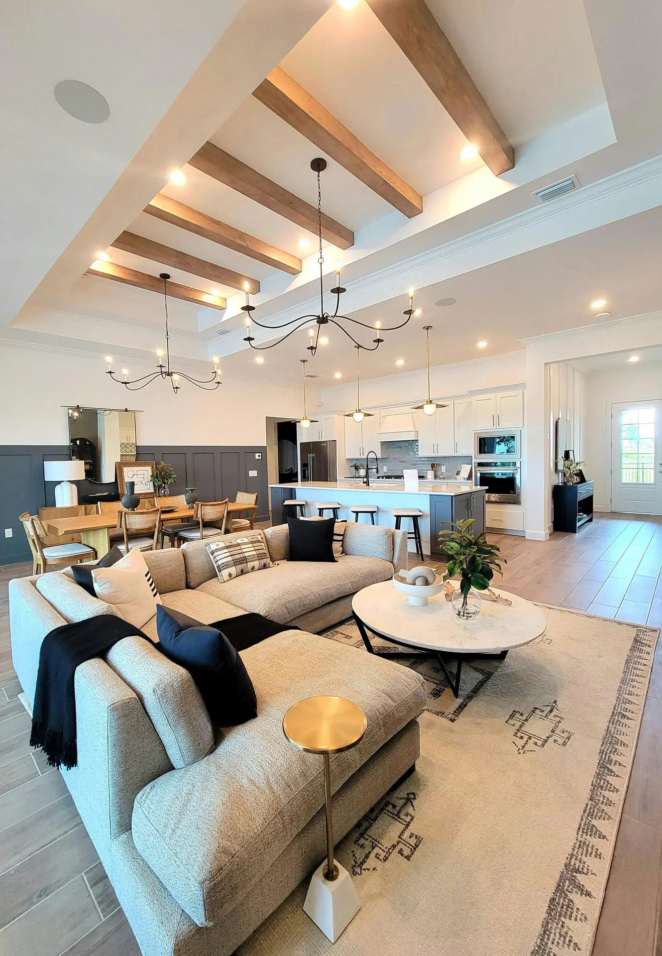 An expansive open-concept great room that masterfully combines coastal living room ideas with Modern Coastal Aesthetics. The living area is anchored by a large, light grey sectional sofa adorned with black and white decorative pillows, centered around a white marble coffee table and a patterned area rug. High coffered ceilings with exposed wooden beams and elegant black wrought-iron chandeliers enhance the sense of space. In the background, a seamless transition leads to a sophisticated dining area with navy blue wainscoting and a gourmet kitchen featuring a large island with bar seating and white cabinetry. Light wood flooring and abundant recessed lighting tie the luxurious, airy space together.