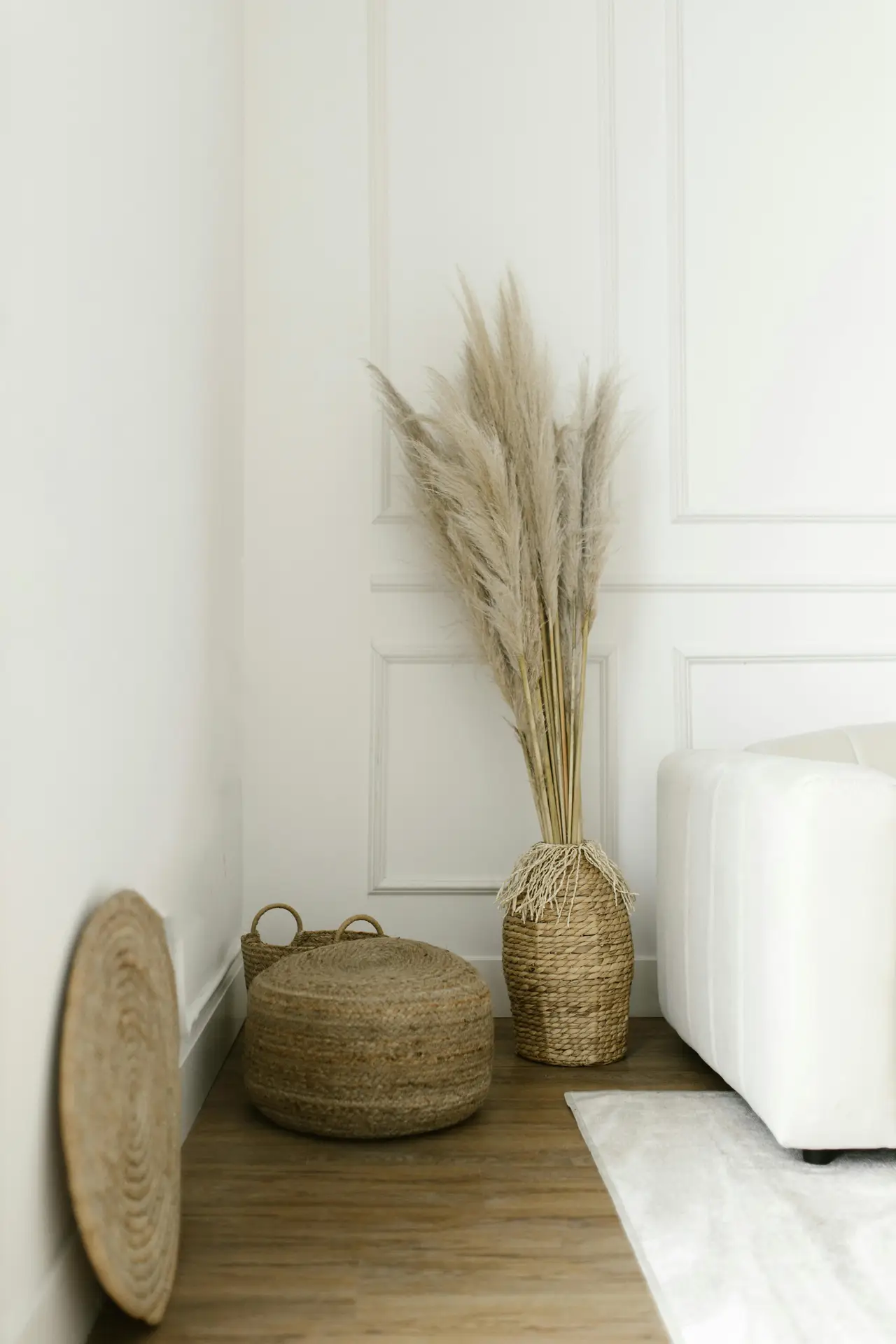 A serene corner of a room that perfectly captures organic coastal minimalism as part of various coastal living room ideas. Against a crisp white paneled wall, a tall woven seagrass vase holds a large, feathery arrangement of dried pampas grass. Beside it, a textured jute pouf and a small woven basket sit on the dark wood floor next to the edge of a modern white sofa. The minimalist composition uses natural fibers and a neutral color palette to create a calm, breezy, and coastal-inspired atmosphere.