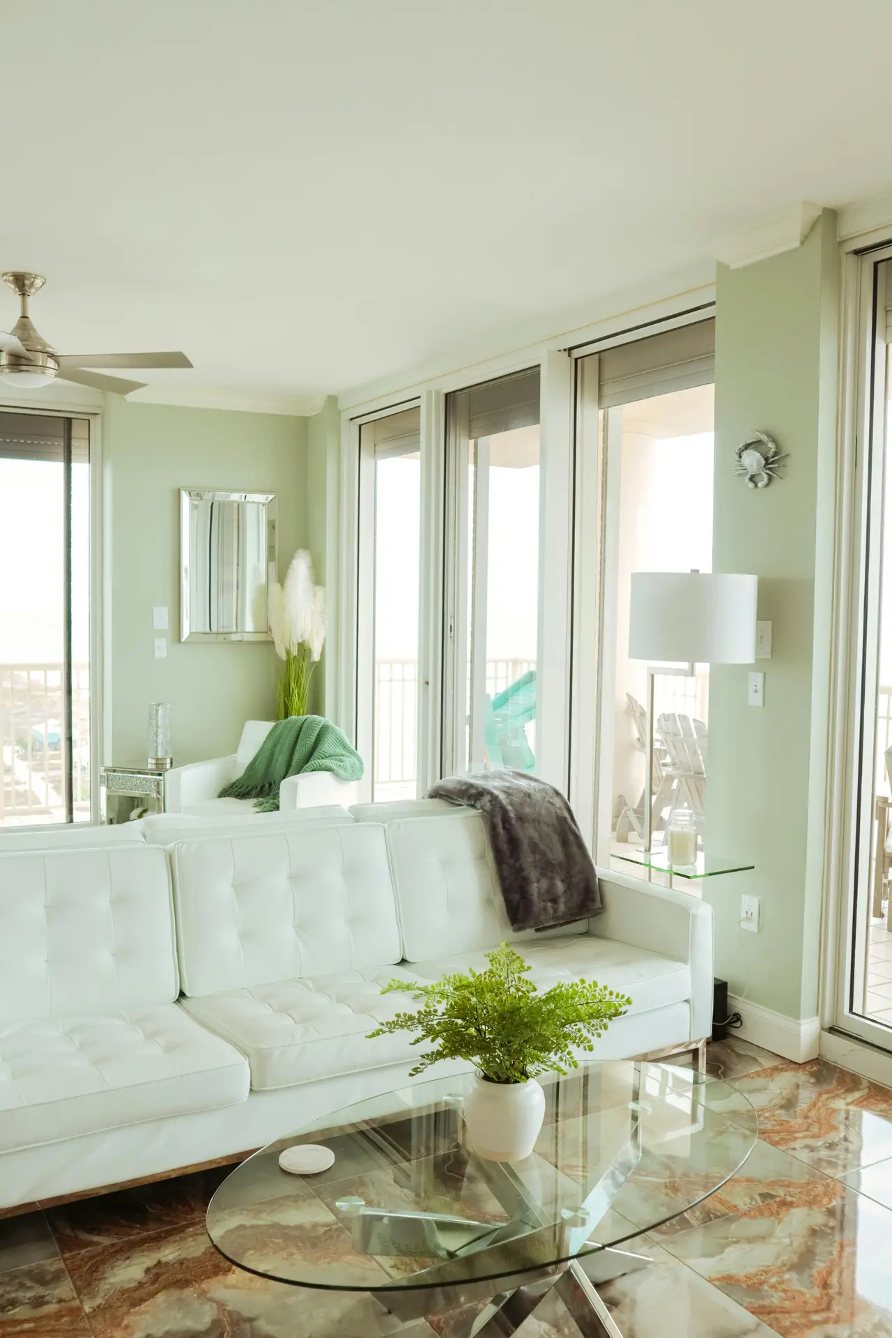 A serene and inviting space illustrating coastal living room ideas with a harmonious sage green and white palette. The scene captures the corner of a sage green sofa, softened by a white and cream patterned pillow and a chunky knit throw blanket. Resting on the wooden armrest are a white coffee mug on a small saucer, a notebook, and a gold pen. The background features a large window draped with sheer white curtains, filtering natural light over potted plants and a woven lamp shade. A textured round jute rug defines the light wood flooring.