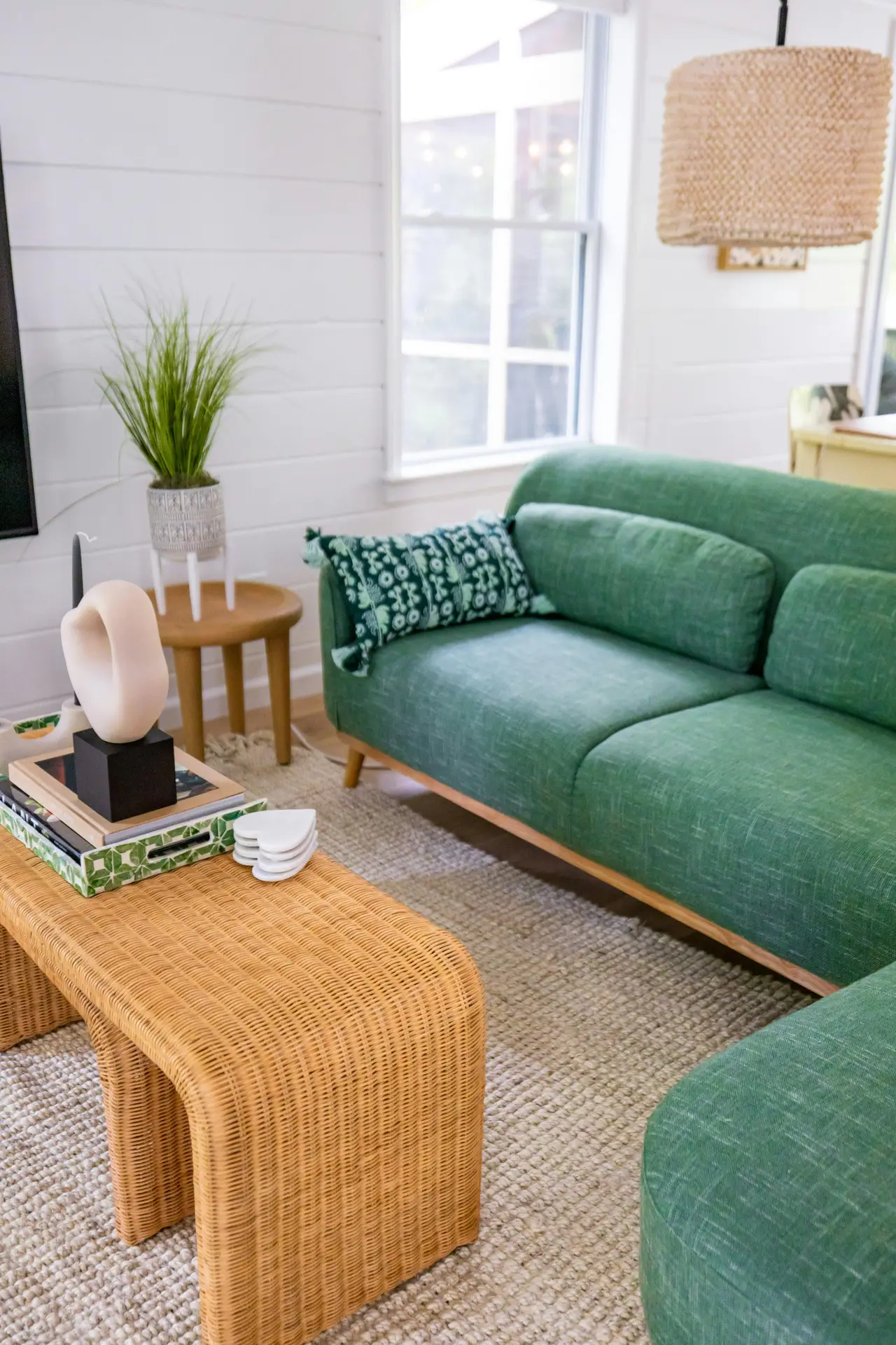 A bright and refreshing space filled with coastal living room ideas. The center of the room features a vibrant Seefoam Green Couch & Rattan Coffee Table set upon a textured neutral rug. The woven coffee table is styled with a decorative tray, books, and a modern white sculpture, while the green sofa is accented with a patterned emerald throw pillow. White shiplap walls, a large window, and a woven pendant light create a light-filled, organic atmosphere, complemented by a small wooden side table holding a green potted plant.