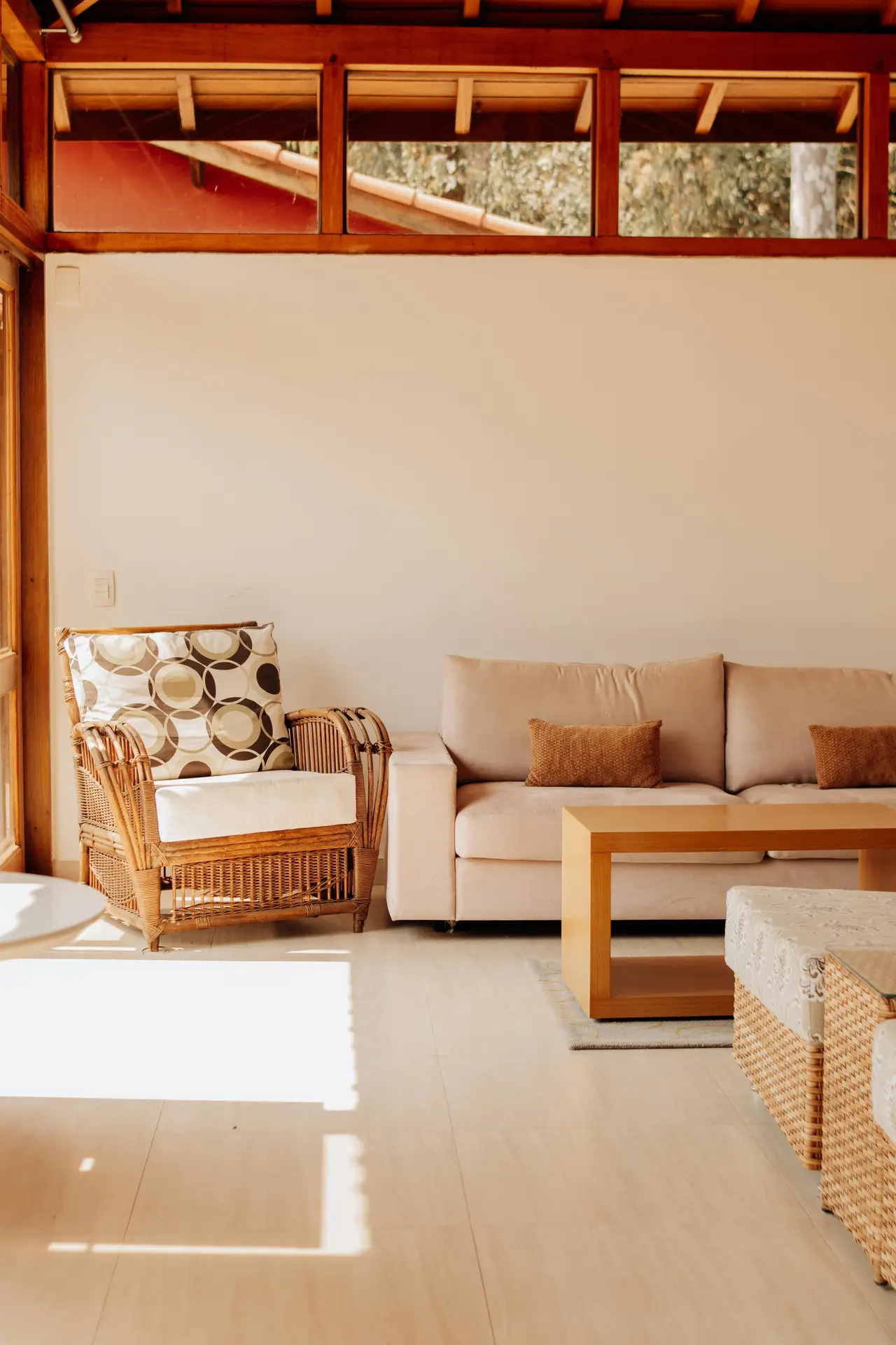 A warm and sun-drenched space that beautifully illustrates coastal living room ideas through a palette of soft sand tones. The room features a comfortable beige sofa accented with cinnamon-colored pillows alongside a classic wicker armchair with a patterned geometric cushion. Natural light streams through a row of high windows set in a dark wood frame, illuminating the pale flooring and a minimalist light wood coffee table. The use of organic textures like rattan and wicker creates a relaxed, earthy atmosphere reminiscent of a tranquil beach house.