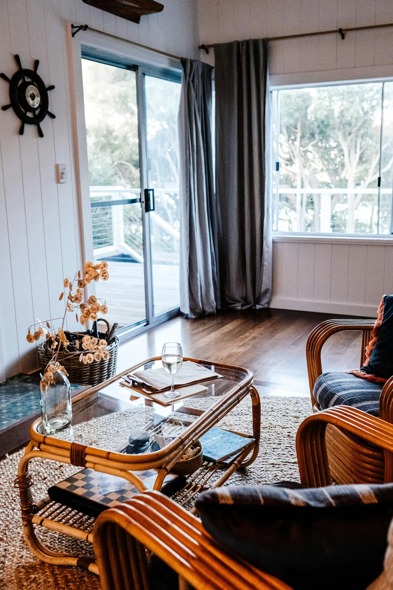 A sun-drenched coastal living room, providing creative coastal living room ideas. A set of vintage rattan furniture, including a sofa and armchair with striped blue and white cushions, sits on a jute rug. Woven pendant lights hang above a glass-top coffee table and another round table. The space is bright and breezy, with natural light streaming through large windows looking out to the ocean and palm trees, creating a relaxed and inviting atmosphere.
