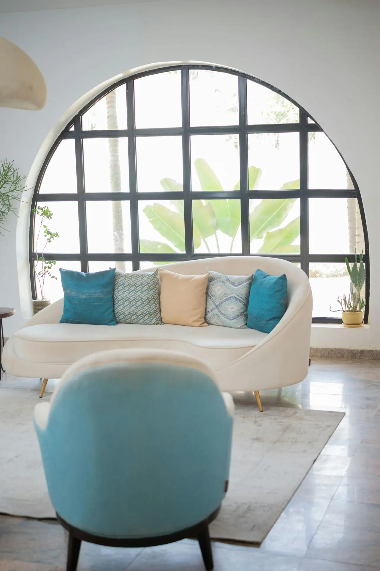 A bright and airy space featuring coastal living room ideas with a harmonious white and blue combo. Sunlight streams through a large window, illuminating a white patterned armchair and a round wooden side table. On the table, a clear glass vase holds green foliage, next to a blue and white ceramic pot, a white candle, and a stack of books with a blue spine. A blue and white woven rug covers the light wood floor, adding a textured touch to the maritime theme.