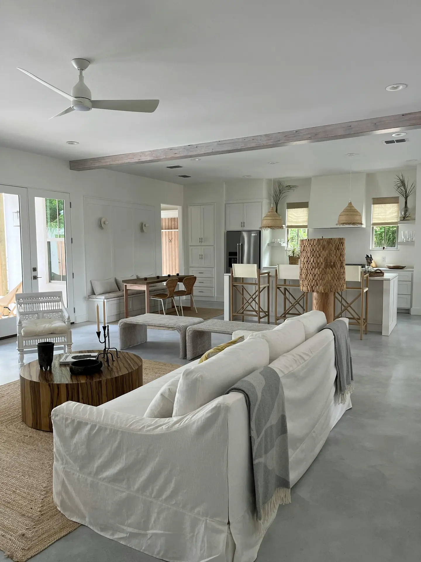A serene and modern interior displaying refined coastal living room ideas. A thick, grey-stained wooden frame mirror is mounted on a textured white shiplap wall above a rustic, reclaimed wood console table. On the table, two white-painted vases with rough textures hold sprigs of preserved baby's breath, next to a lit candle on a small ceramic tray. Woven baskets are nestled in the open shelf below, enhancing the white, grey and wood aesthetic with natural textures.