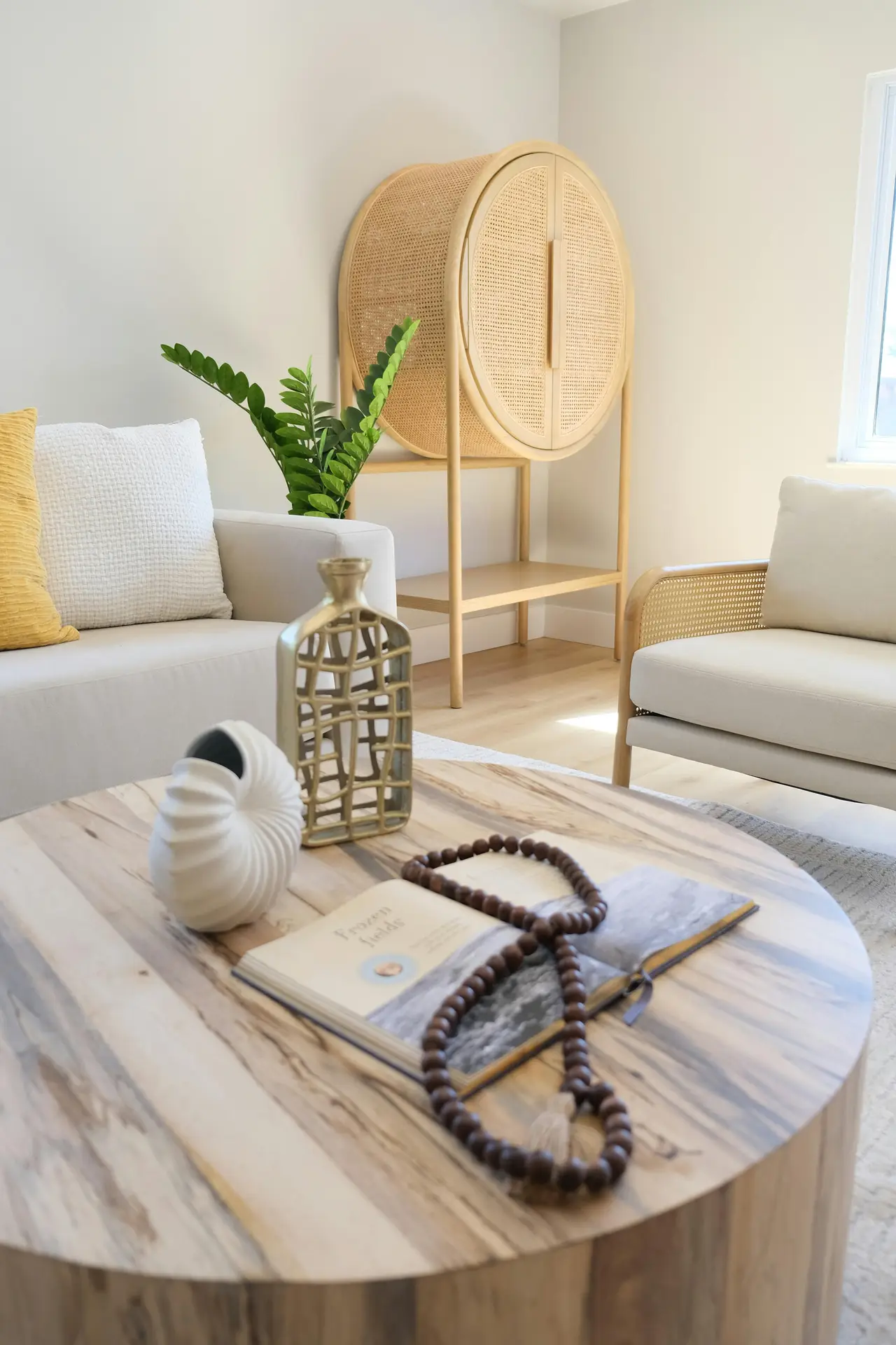 A bright, contemporary interior showcasing inspired coastal living room ideas. In the foreground, a round light-wood coffee table is decorated with an open coffee table book draped with dark wooden beads and shell vase in a stylized nautilus shape. The background features a minimalist cream sofa and a matching armchair with rattan sides. A unique, circular light-wood cabinet with mesh doors stands against the neutral wall next to a vibrant green plant, creating a clean and breezy seaside atmosphere.