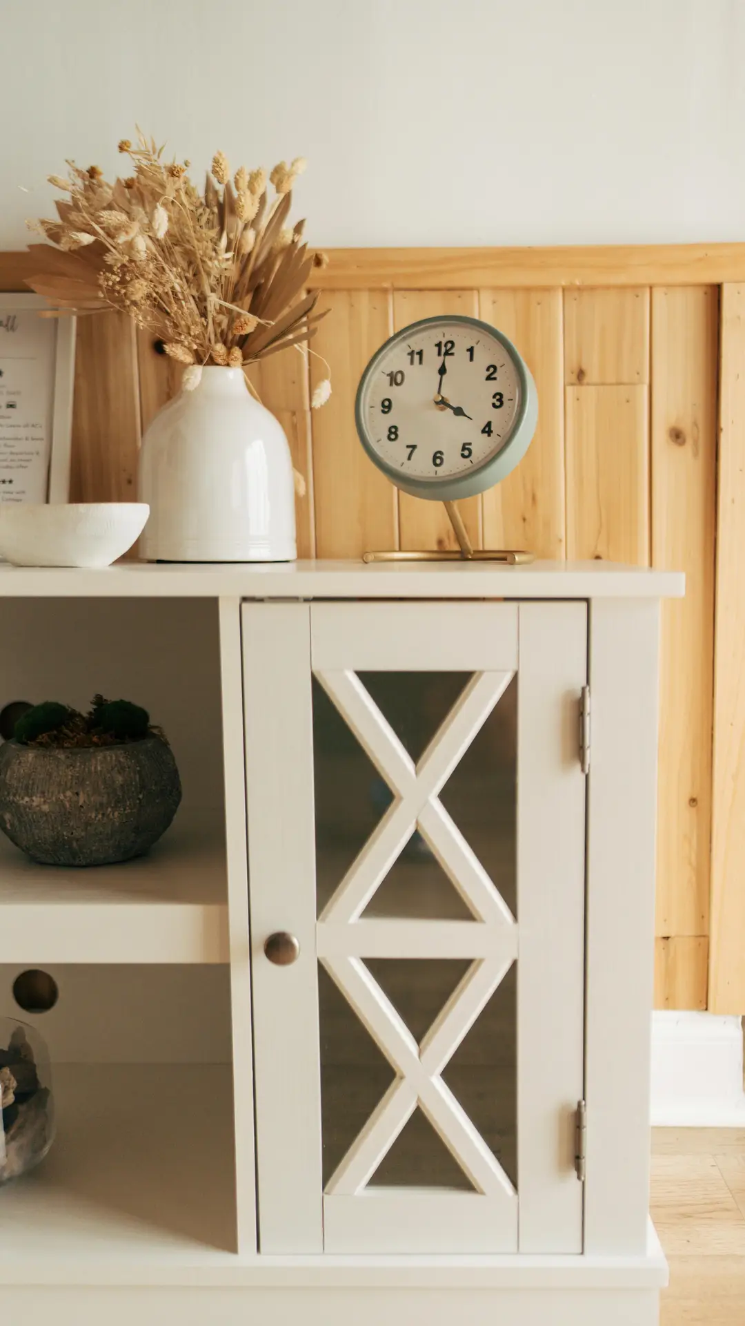 A warm and inviting living area illustrating coastal living room ideas with a refined rustic touch. A large, beautifully textured wooden cupboard and dry flowers serve as the central focus. The cabinet features intricate carved panels and is styled with various decorative items. Atop it rests a large white-painted vase holding a massive, sprawling arrangement of tall pampas grass and other dried botanicals. To its right are several framed pictures, a small lit lantern, and more small, leafy dry flowers. Natural light streams in from a window on the far left, illuminating the soft, sandy tones of the walls and the jute rug below. In the corner to the right, a comfortable cream-colored armchair is placed next to a floor lamp, creating a cozy reading nook.